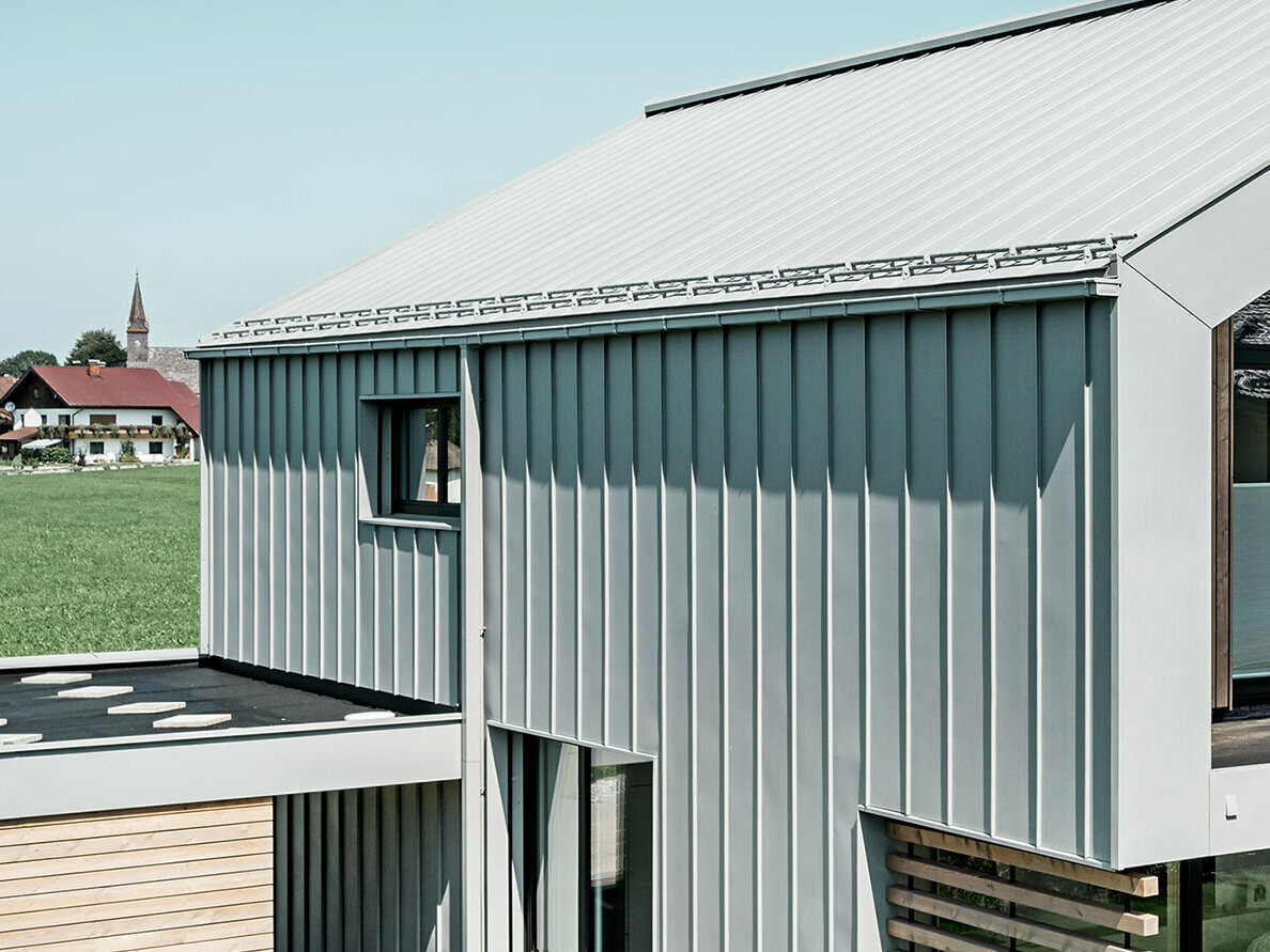 Enodružinska hiša z dvokapno streho, z trakovi PREFALZ na strehi in fasadi; za odvodnjavanje strehe s pravokotnim žlebom PREFA s pravokotno iztočno cevjo PREFA v posebni patinirano sivi barvi.