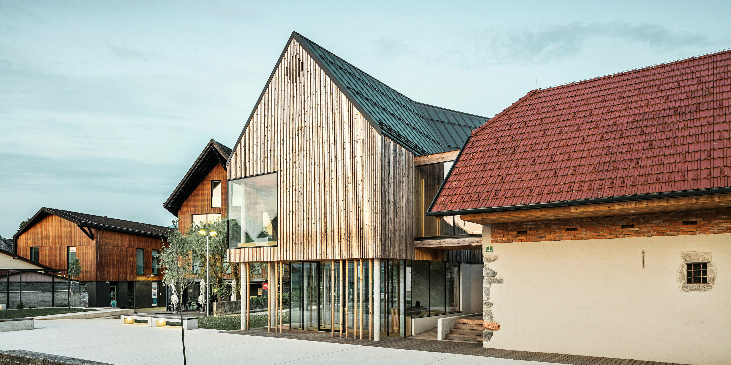Pogled na center za obiskovalce na Igu v Sloveniji z vpadljivo streho PREFALZ v  P.10 temno sivi barvi, ki jasno izstopa iz okoliške tradicionalne rdeče strešne kritine. Kombinacija starega in novega, ki jo predstavljata sosednji zid z majhnim oknom in sodobna lesena konstrukcija z velikimi steklenimi površinami, priča o premišljeni simbiozi regionalne tradicije in sodobne arhitekture. Elegantna osvetlitev poudarja vabljivo vzdušje vhodnega prostora, kot je predstavljeno v knjigi PREFARENZEN 2023.