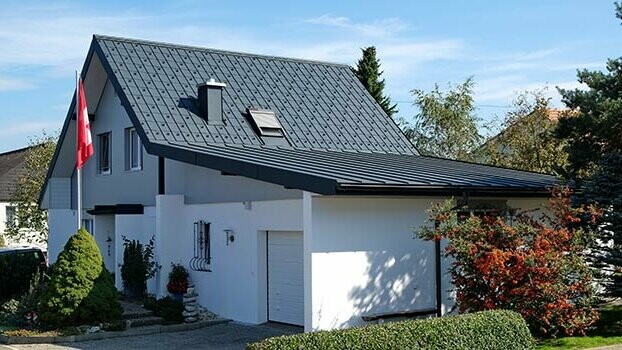 Sanirana hiša z dvokapno streho in priključeno garažo. Hišna streha je prekrita s strešnimi ploščami PREFA, garaža pa z elementi PREFA Prefalz v antracitni barvi. Pred hišo stoji steber s švicarsko zastavo.