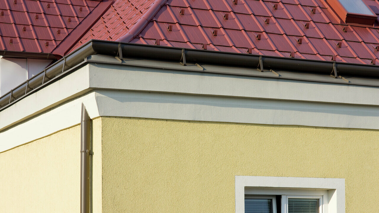 Vogal hiše z rumeno ometano fasado in rjavim ležečim žlebom PREFA (strešni žleb) s streho z rdečimi strešnimi ploščami PREFA