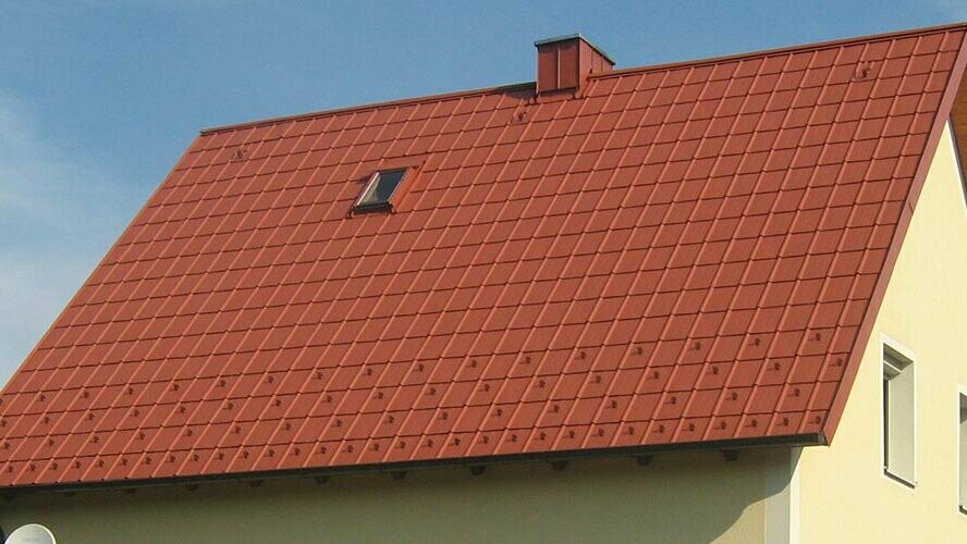 Klasična dvokapna streha z majhnim strešnim oknom in dimnikom, prekrita s PREFA strešno ploščo v opečno rdeči barvi, vključno s snegolovi in varnostnimi kavlji.