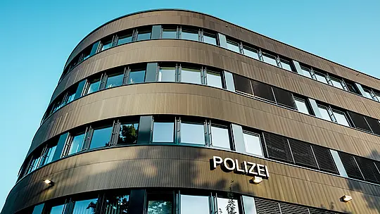Dramatičen pogled od spodaj na ukrivljeno novo stavbo policijske postaje 43 v Hamburgu: izrazita PREFA aluminijasta fasada iz eloksiranih ekstrudiranih profilov »zobčasti profil« v srednje bronasti barvi privlači vse poglede kot arhitekturni magnet. Fina, ritmična struktura ustvarja dinamične prehode svetlobe in sence ter poudarja tekoče zaobljenosti stavbe. PREFA aluminij prepriča z visoko odpornostjo proti vremenskim vplivom, zaščito pred korozijo in dolgo življenjsko dobo – trajnostna rešitev, ki zahteva malo vzdrževanja, za natančno, moderno fasadno arhitekturo. 