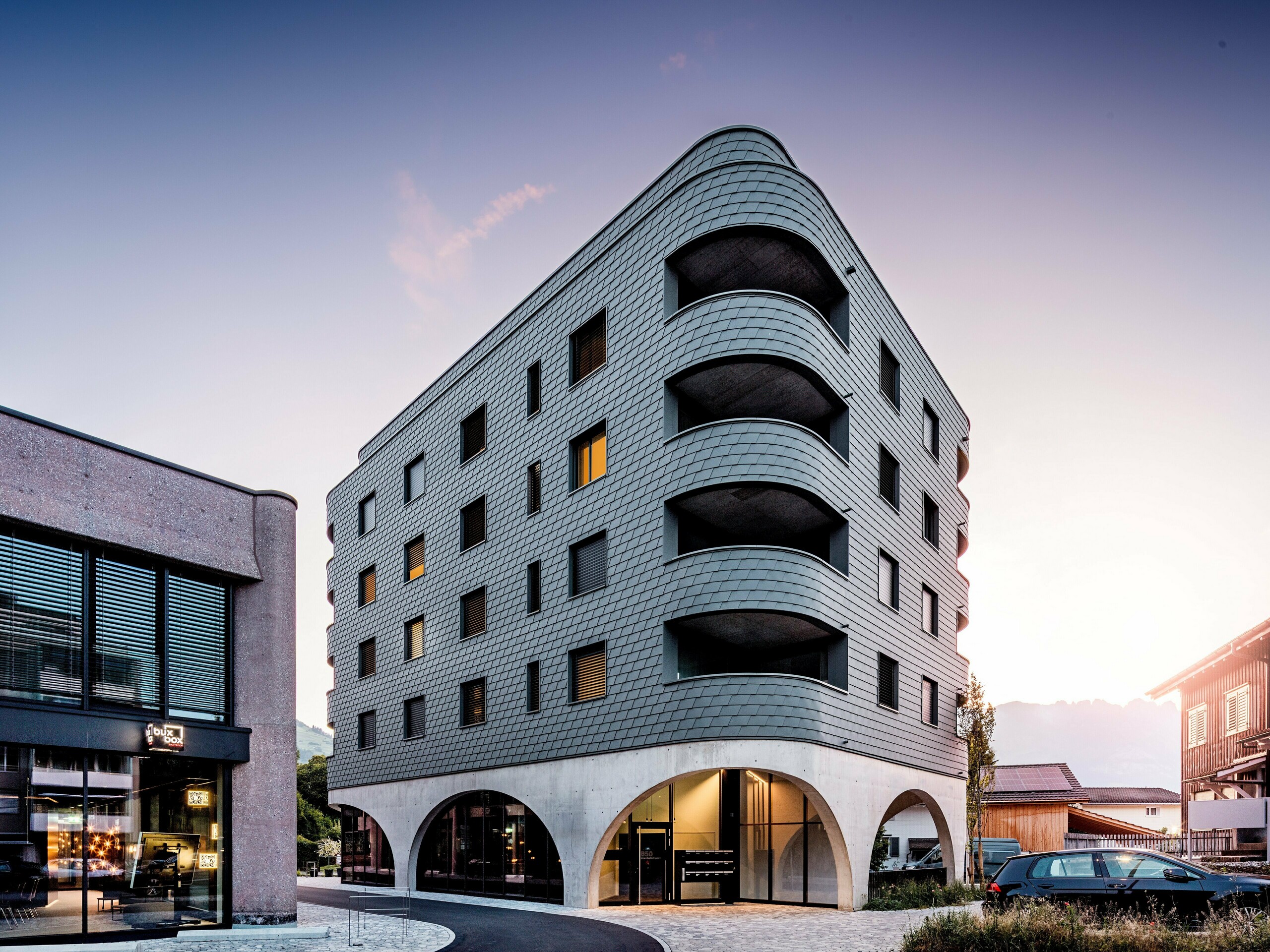 Sodobna večnamenska stavba v Buchsu z izrazito zaobljeno, trikotno obliko in neprekinjeno fasado iz PREFA fasadnih skodel v barvi P.10 svetlo siva. Luskasta aluminijasta obloga ovija stavbo kot struktura plazilca in poudarja tekoče zaobljenosti ter globoko vrezane balkone. Nad masivnim podstavkom z elegantnimi betonskimi arkadami deluje lahka, vremensko odporna fasada PREFA še posebej izrazito. Natančno položene aluminijaste skodle zagotavljajo homogeno, trajno in vzdrževanju prijazno ovojnico stavbe z močno prepoznavnostjo.