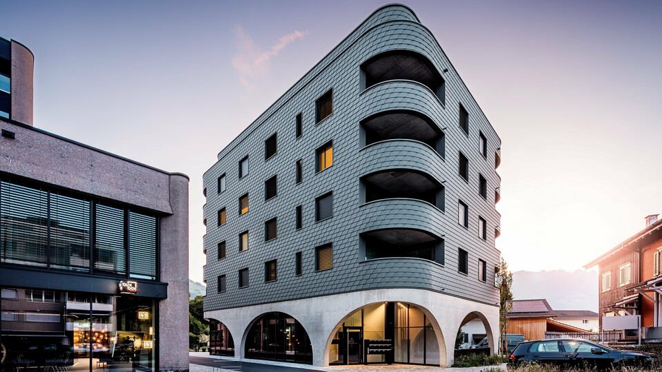 Sodobna večnamenska stavba v Buchsu z izrazito zaobljeno, trikotno obliko in neprekinjeno fasado iz PREFA fasadnih skodel v barvi P.10 svetlo siva. Luskasta aluminijasta obloga ovija stavbo kot struktura plazilca in poudarja tekoče zaobljenosti ter globoko vrezane balkone. Nad masivnim podstavkom z elegantnimi betonskimi arkadami deluje lahka, vremensko odporna fasada PREFA še posebej izrazito. Natančno položene aluminijaste skodle zagotavljajo homogeno, trajno in vzdrževanju prijazno ovojnico stavbe z močno prepoznavnostjo.