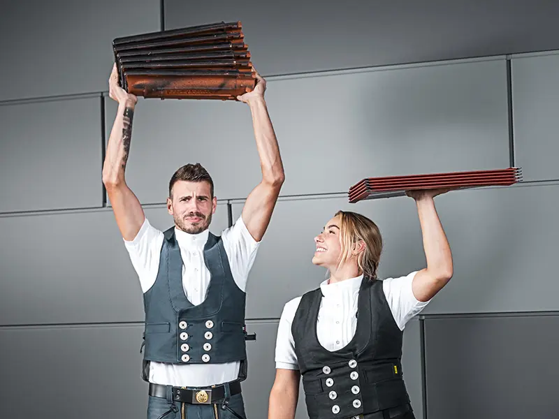 Na sliki sta prikazani dve osebi, ki v rokah držita kup strešnega materiala. Oseba na levi strani drži kup strešnikov nad glavo, medtem ko oseba na desni strani drži kup aluminijastih strešnih plošč prav tako nad glavo. Oba nosita tradicionalna oblačila za obrtnike z jopico in belo srajco. Ozadje je moderno in minimalistično, s sivimi stenami. Obe osebi delujeta osredotočeni in se smehljata.