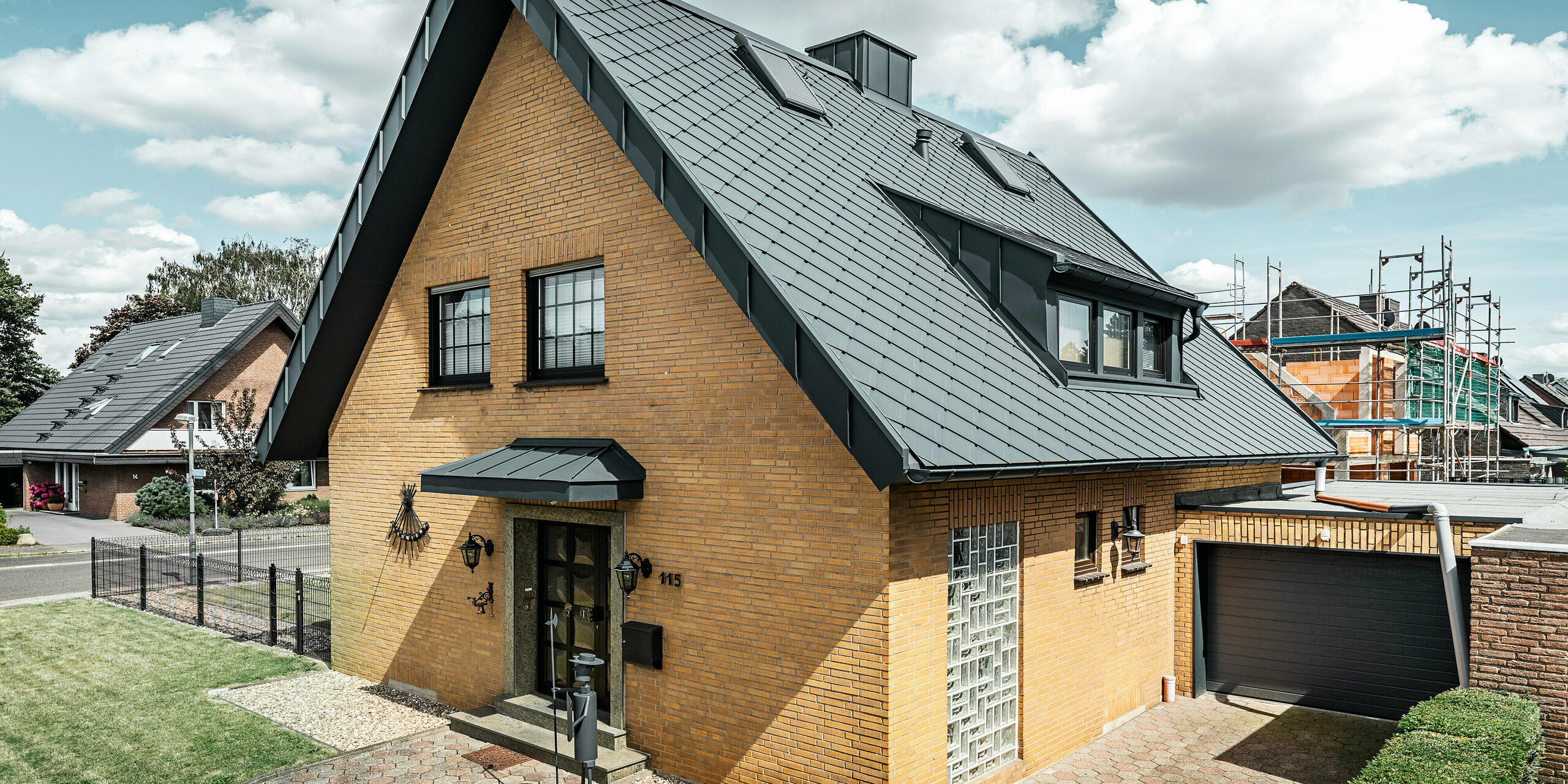 Vhodni del tradicionalne enodružinske hiše z moderno PREFA aluminijasto streho s strešnimi rombi 29 × 29 v P.10 antracitni barvi. Strešna kritina vhodnih vrat, mansarde in izstopajoči dimnik so prekriti s sistemom PREFALZ. Enotno strešno pokrajino zaokrožujeta viseči žleb in odtočna cev, tudi v barvi P.10 antracit. Hiša stoji v bližini manjšega mesta. V ozadju so vidni gradbeni odri s sosednjega gradbišča. Gradbeni oder štrli neposredno nad sosednjo garažo, ki je prav tako zidana.
