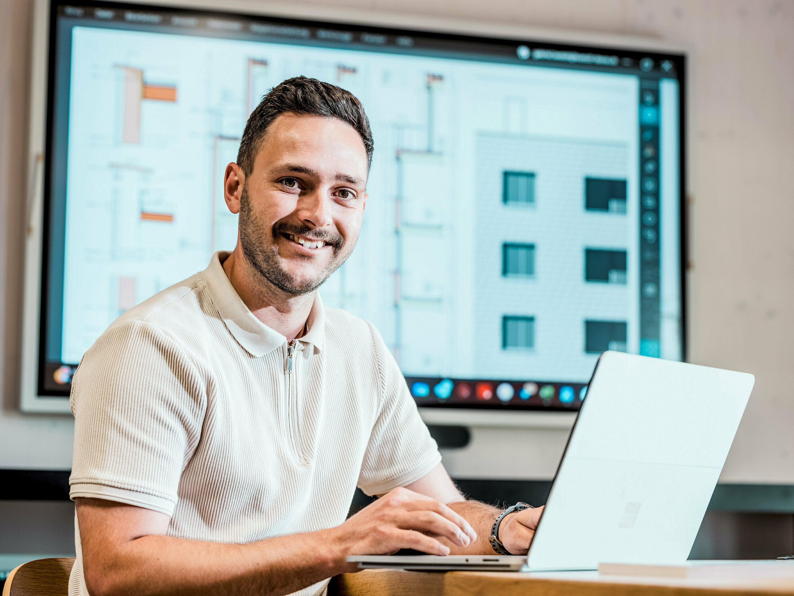 Portret Gabriela Landolta, zaposlenega v podjetju Künzli Davos AG, za prenosnim računalnikom pred velikim zaslonom, na katerem so vidni digitalni gradbeni načrti in podrobnosti fasade. Na zaslonu so vidne tehnične risbe in pogledi na moderno stavbo s fasado v lusksasti obliki, ki je bila izvedena s PREFA fasadnimi skodlami v barvi P.10 svetlo siva. Inovativna aluminijasta fasada prepriča s svojo nizko težo, odpornostjo proti vremenskim vplivom in dolgo življenjsko dobo. Natančno načrtovanje in uporaba visokokakovostnih izdelkov PREFA sta osnova za oblikovno stabilno, vzdrževanju prijazno ovojnico stavbe z arhitekturnimi zahtevami.