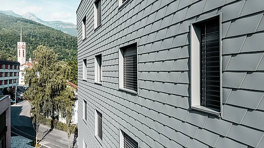 Podrobni pogled na fasado hiše Bernerhaus v Buchsu z natančno položenimi PREFA fasadnimi skodlami v barvi P.10 svetlo siva. Luskasta aluminijasta površina ustvarja fin, živahen vzorec in poudarja jasno izrezane okenske odprtine z globokimi okenskimi podboji. Lahki, vremensko odporni PREFA aluminij zagotavlja homogeno, oblikovno stabilno in vzdrževanju prijazno ovojnico stavbe. Pred alpskim ozadjem postane visokokakovostna kovinska fasada sodoben magnet za poglede s brezčasnim izžarevanjem.