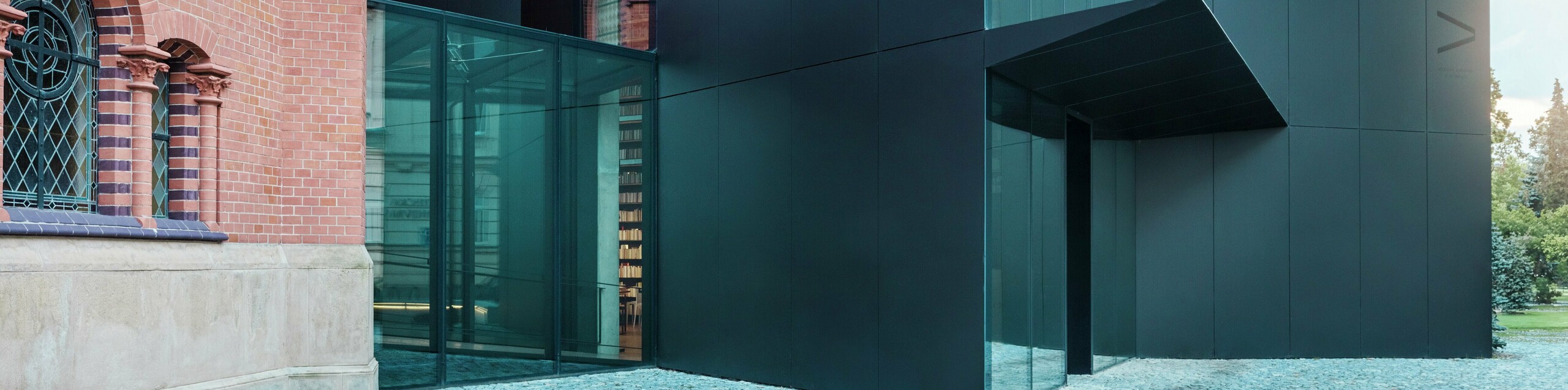 Slika prikazuje moderno razširitev knjižnice, ki se elegantno navezuje na zgodovinsko Rdečo cerkev v Olomouncu. Izrazita nova stavba navdušuje s fasado iz črnih PREFABOND aluminijastih kompozitnih plošč, ki je oblikovana v jasnih, geometrijskih oblikah in prepriča s precizno izdelanimi detajli. Steklene stene stavbe omogočajo prosojen pogled v notranjost, ki se harmonično vklaplja v zgodovinsko okolico. Sodelovanje sodobnih materialov in klasičnih arhitekturnih elementov ustvarja zanimivo vizualno povezavo med starim in novim. Inovativna uporaba plošč PREFABOND ne zagotavlja le sodobnega videza, ampak tudi dolgo življenjsko dobo in odpornost, ki stavbo ščitita za prihodnost.