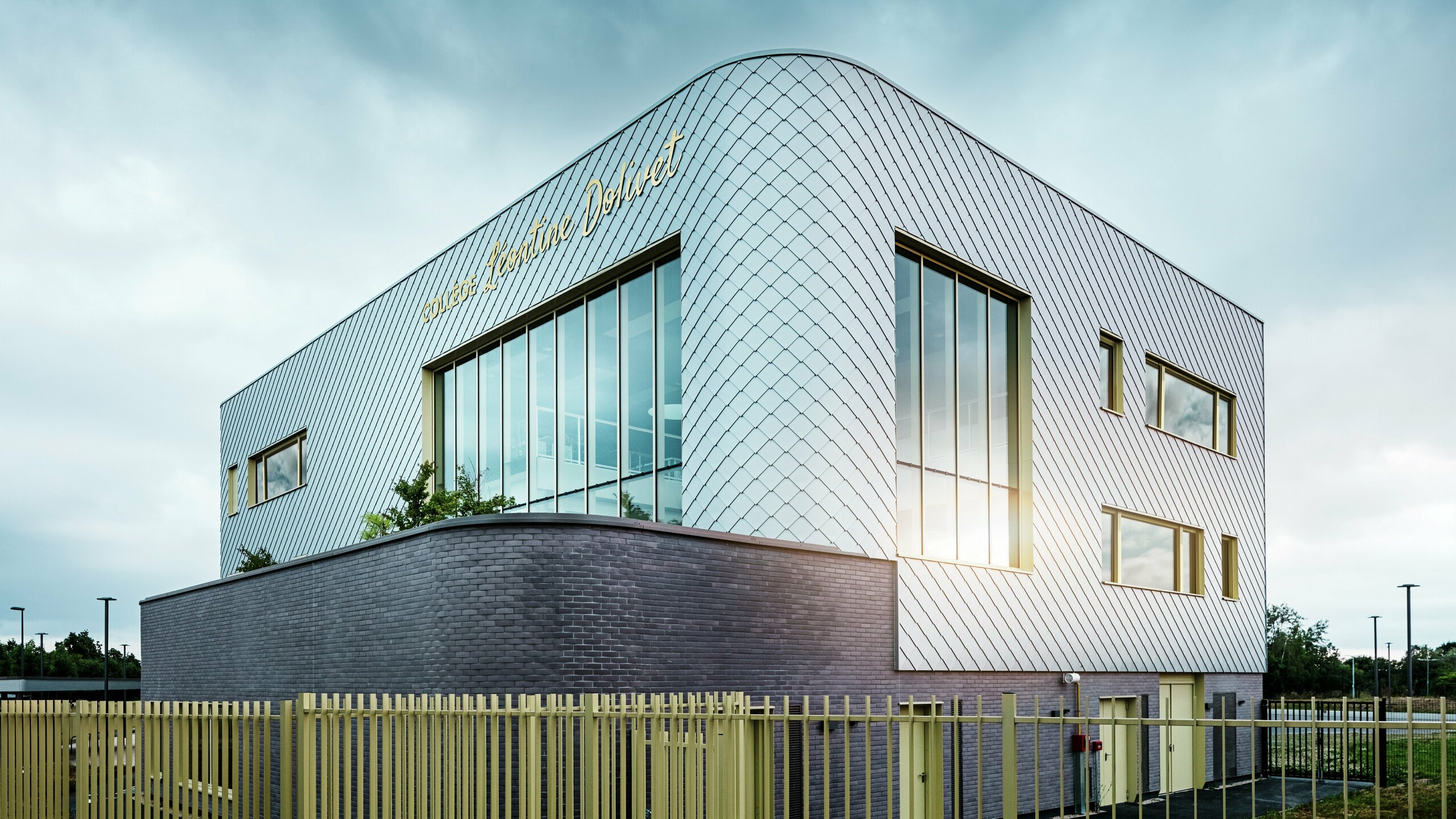 Fotografija prikazuje markanten kot Collège Léontine Dolivet z ukrivljeno aluminijasto fasado iz kovinsko srebrnih PREFA fasadnih rombov. Velike steklene površine odbijajo svetlobo, rumeni okenski okvirji pa dodajajo poudarke. V ospredju se vzdolž stavbe razteza svetla ograja, ki stoji na temnem podstavku iz opeke. Nebo je oblačno.