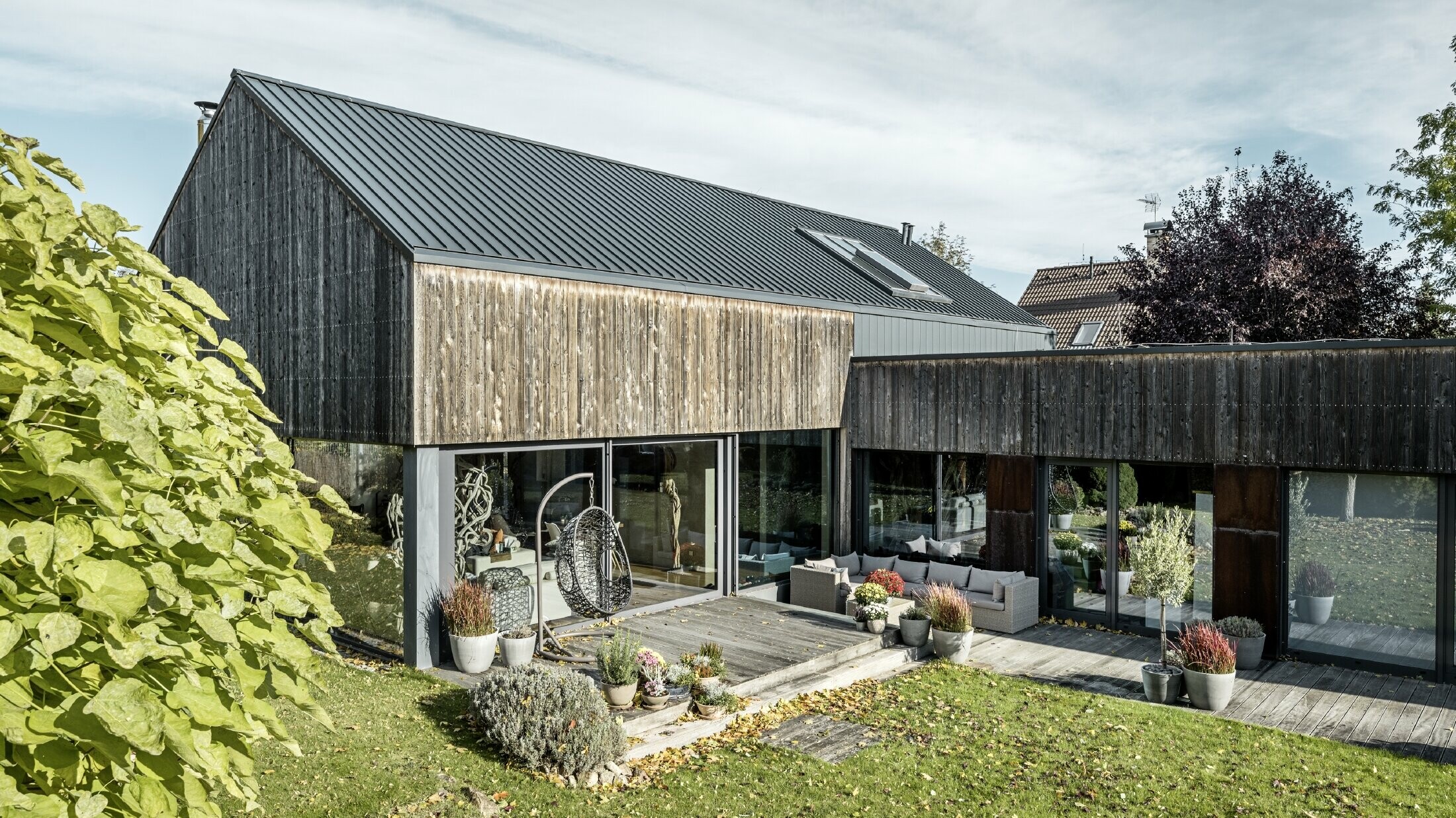Enodružinska hiša z dvokapno streho, obložena z dvojnimi stoječimi zgibi PREFALZ v antracitni barvi in z leseno fasado. Z lepo leseno teraso in velikimi okenskimi površinami v pritličju.