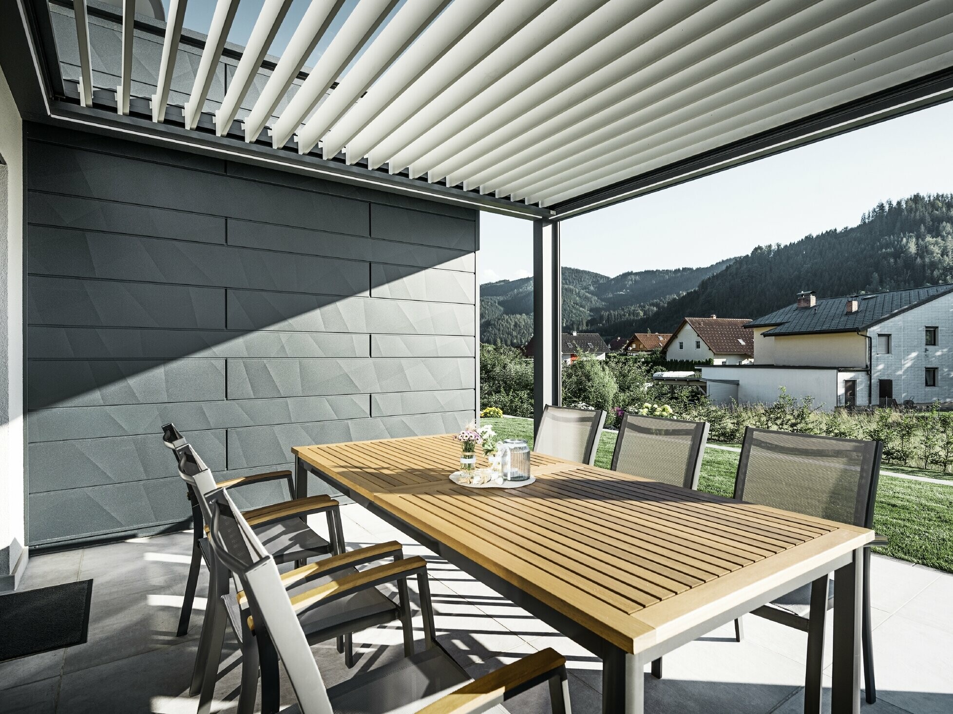 Z lamelami pokrita, zaščitena terasa z mizo in stoli; ena stran fasade je obložena s PREFA Siding.X fasadnimi paneli v antracitni barvi.