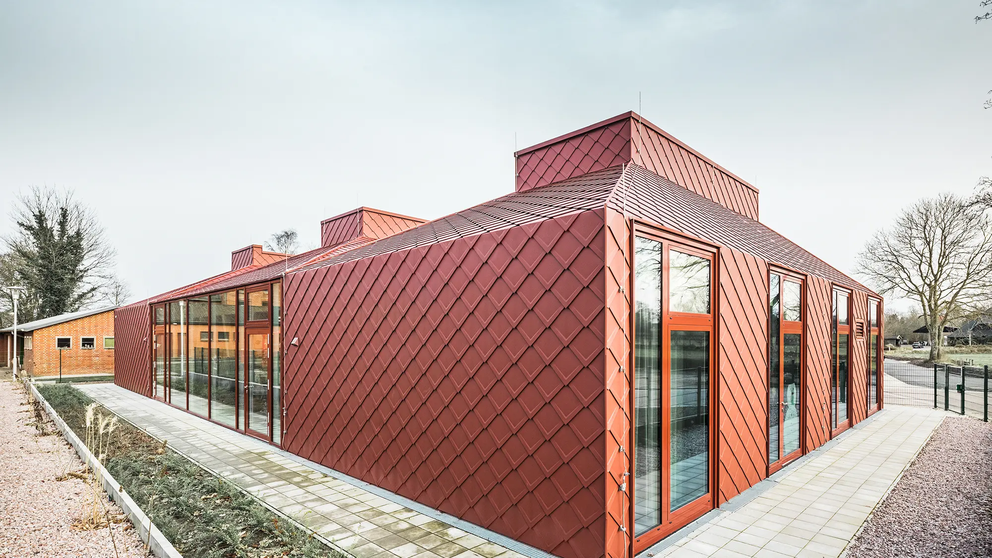 Ein modernes, einstöckiges Gebäude mit einer roten Fassade aus Aluminium, gedeckt mit PREFA Dach- und Wandrauten. Zu sehen sind große, raumhohe Fensterfronten sowie einer klaren, modernen Architektur, fotografiert bei Tageslicht in einer Frontalaufnahme.