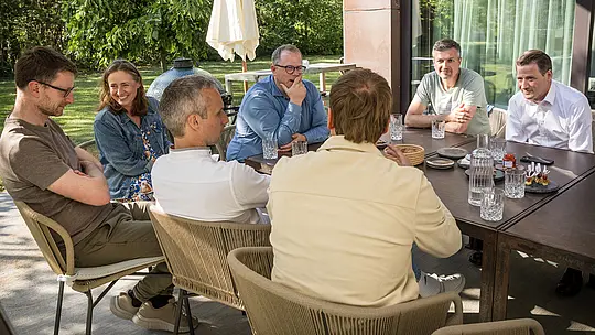 PREFARENZEN 2027 Strokovni dialog v soncu prežetem zunanjem prostoru vinarske rezidence: arhitekti in strokovnjaki za oblikovanje sedijo okrog velike mize, globoko zatopljeni v razprave, medtem ko so na mizi razporejeni kozarci in majhni prigrizki. Prizor prikazuje živahno interakcijo in osredotočeno pozornost, obdano z naravno zeleno barvo in senco dreves. PREFA aluminijasti izdelki v ozadju predstavljajo trajne, estetsko privlačne strehe in fasade. Slika prikazuje, kako PREFA združuje oblikovanje, funkcionalnost in trajnost ter navdihuje kreativne razprave med arhitekti.