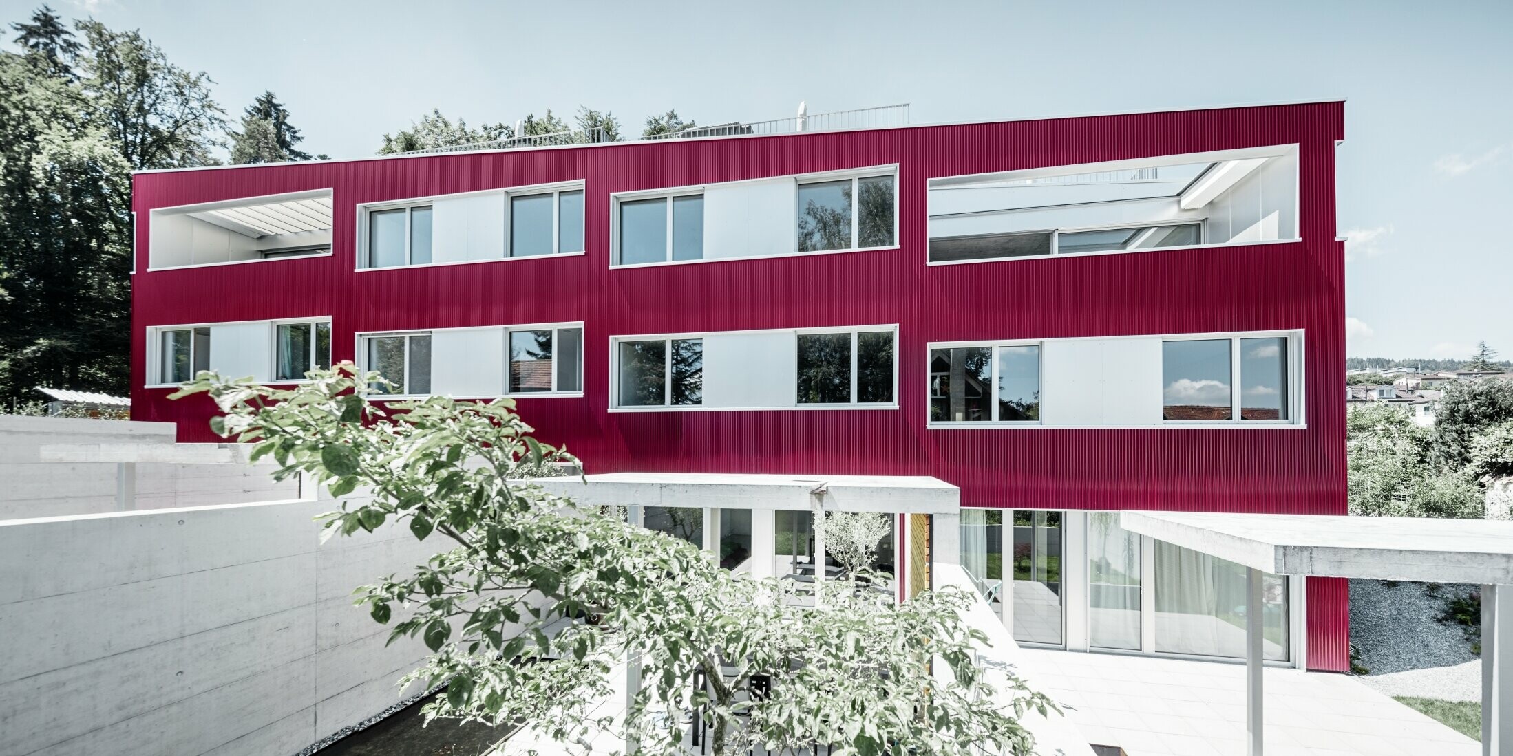 Večstanovanjski objekt z ravno streho in balkoni s fasado vinsko rdeče barve z valovitim profilom PREFA 