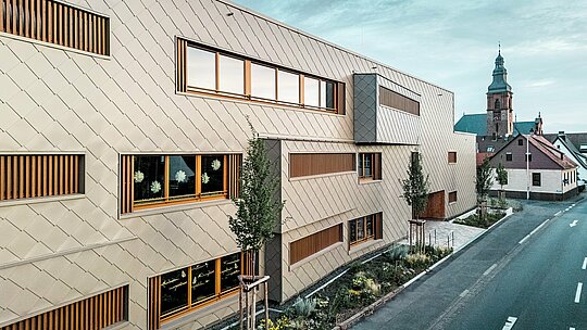 Stranski pogled na vrtec v mestu Alzenau prikazuje moderno hibridno stavbo v zanimivem kontrastu z zgodovinsko mestno strukturo in poznogotsko cerkvijo v ozadju. Fasada iz bronastih PREFA fasadnih rombov 44 × 44 ustvarja eleganten, ritmičen vzorec in se samozavestno loči od okolice, ne da bi bila vsiljiva. Prostorne okenske odprtine z navpičnimi lesenimi lamelami in strukturirano zelenje vzdolž stavbe poudarjajo trajnostni koncept oblikovanja. Kombinacija inovativne aluminijaste fasade, ekološke gradnje in urbanistične občutljivosti naredi projekt za vzoren primer odgovorne arhitekture med tradicijo in prihodnostjo.