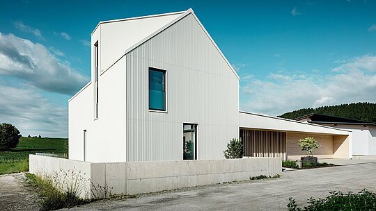 Moderna enodružinska hiša v Freistadtu iz dinamične perspektive, ki poudarja jasne linije in opazno arhitekturo. Bela PREFALZ-aluminijasta obloga se neprekinjeno razteza po celotni fasadi in strehi ter hiši daje moderno in brezčasno estetiko. Značilni elementi zasnove so strma dvokapna streha, velika okna in izrazita strešna okna, medtem ko navpični žlebovi na fasadi stavbi dajejo zanimivo teksturo in globino. Kontrast z naravnim okoljem in elegantni leseni elementi v vhodnem prostoru in na garažnih vratih dopolnjujejo dizajn. PREFALZ zagotavlja posebej trajno, enostavno vzdrževano in vremensko odporno površino. Tako je hiša ne le estetsko privlačna, ampak tudi funkcionalna in trajnostna.