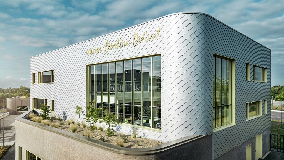 Fotografija prikazuje markanten vogal stavbe Collège Léontine Dolivet v Cesson-Sévigné. Fasada je sestavljena iz kovinsko srebrnih PREFA fasadnih rombov, ki so razporejeni v diagonalnem vzorcu in odbijajo svetlobo. Opazna je ukrivljena oblika zgornjega nadstropja, ki stavbi daje mehak, sodoben videz. Velika steklena fasada omogoča pogled v notranjost, rumeni okenski okviri pa dodajajo živahne poudarke. V ospredju je zasajeno območje s prodnatim tlakom in grmičevjem, ki harmonično dopolnjuje arhitekturo. Nebo je rahlo oblačno, kar poudarja kovinsko površino. 