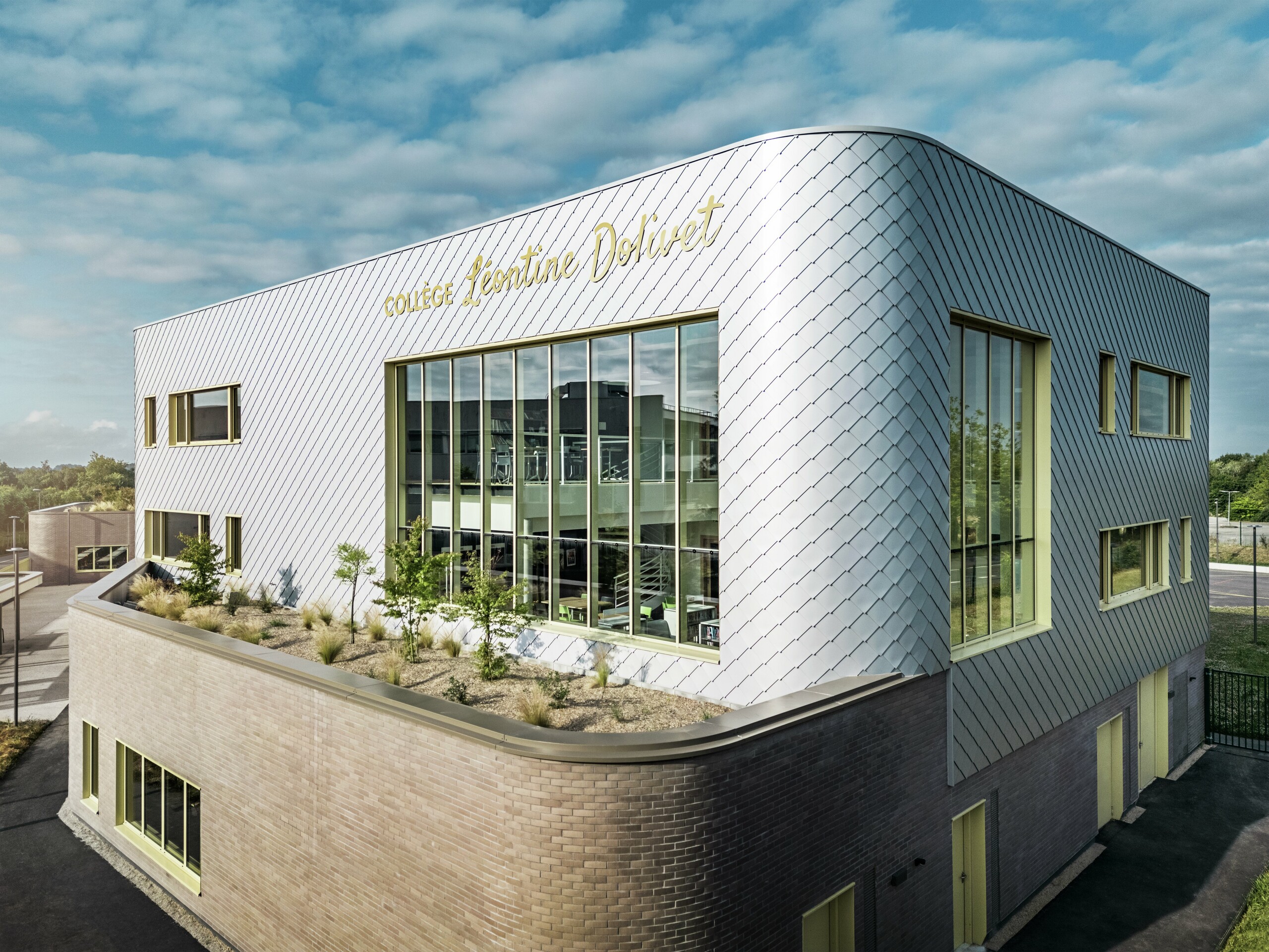 Fotografija prikazuje markanten vogal stavbe Collège Léontine Dolivet v Cesson-Sévigné. Fasada je sestavljena iz kovinsko srebrnih PREFA fasadnih rombov, ki so razporejeni v diagonalnem vzorcu in odbijajo svetlobo. Opazna je ukrivljena oblika zgornjega nadstropja, ki stavbi daje mehak, sodoben videz. Velika steklena fasada omogoča pogled v notranjost, rumeni okenski okviri pa dodajajo živahne poudarke. V ospredju je zasajeno območje s prodnatim tlakom in grmičevjem, ki harmonično dopolnjuje arhitekturo. Nebo je rahlo oblačno, kar poudarja kovinsko površino. 