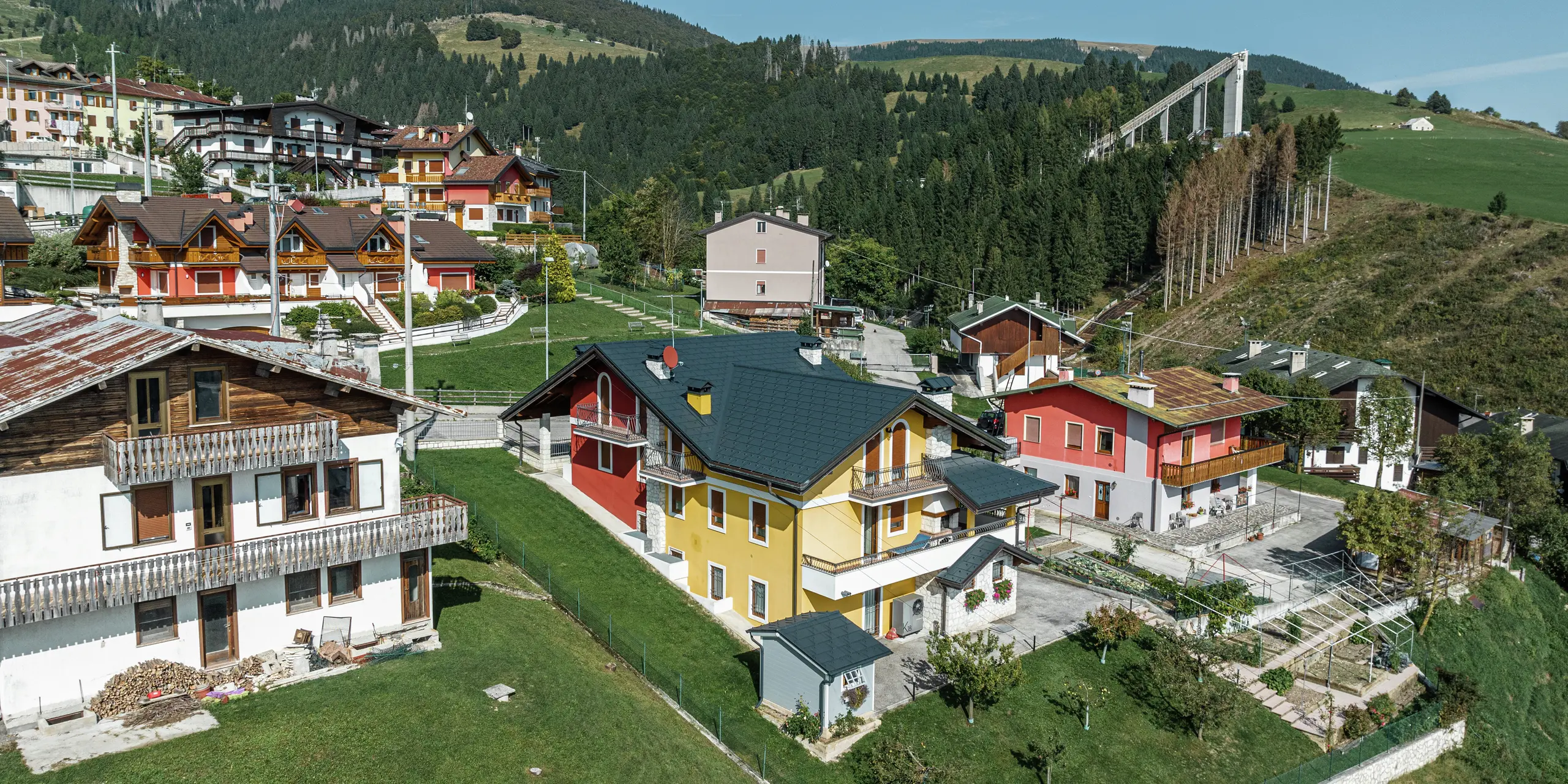Idyllisches Einfamilienhaus in Gallio mit modernem PREFA Blechdach aus Dachplatten R.16 in P.10 Dunkelgrau. Die widerstandsfähige Dacheindeckung aus Aluminium schützt das Haus zuverlässig vor Schnee, Regen und Wind - ideal für alpine Regionen. Die gelb-rote Fassade mit Natursteinakzenten fügt sich harmonisch in die grüne Umgebung ein. Dichte Wälder und eine Skisprungschanze im Hintergrund unterstreichen den typisch alpinen Charakter der Region. Langlebig, robust und ästhetisch - ein perfektes Dach für Generationen.