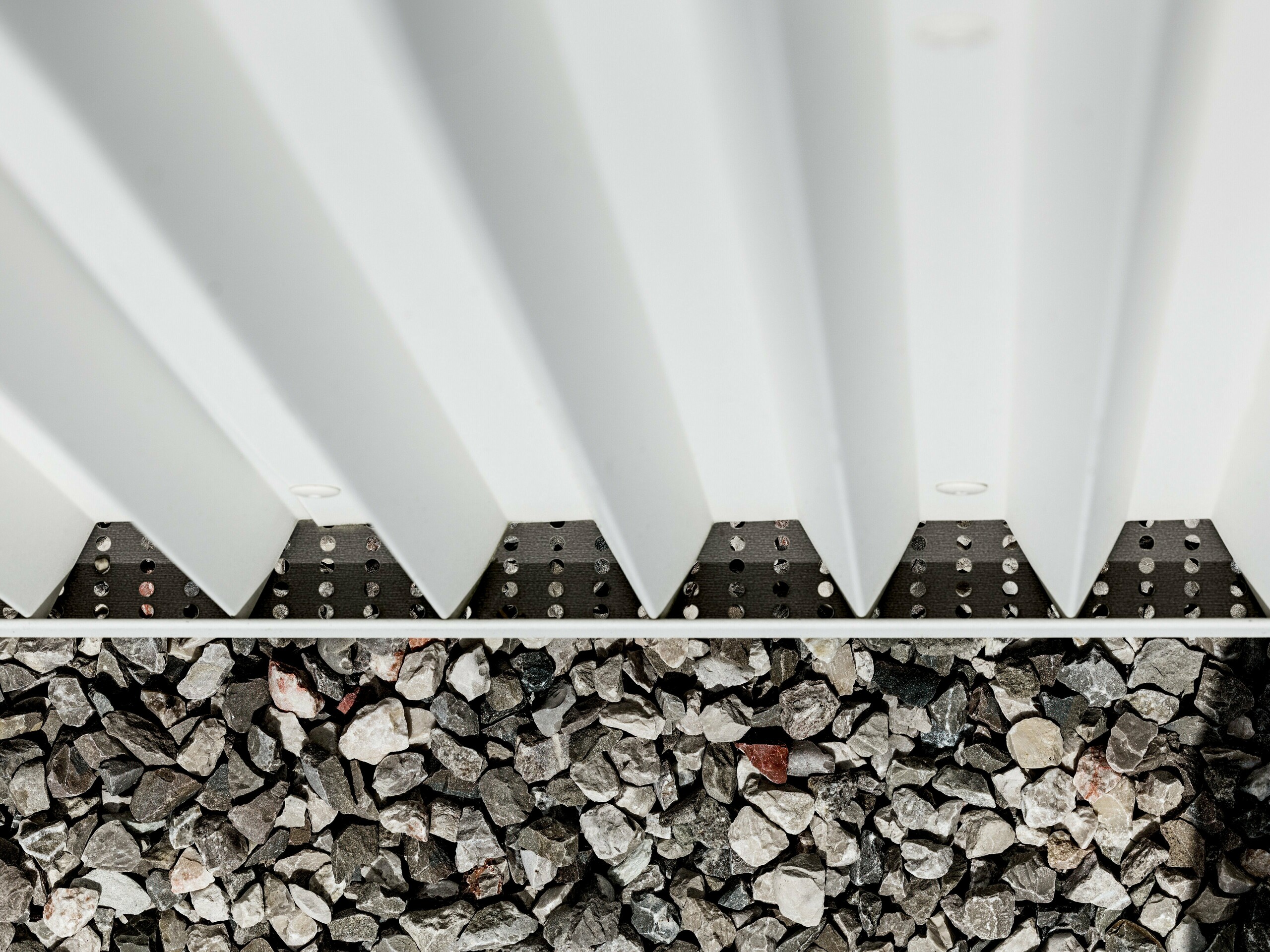 Slika prikazuje podrobnosti prezračevane fasade PREFALZ iz belega aluminija, ki je opremljena s precizno zloženimi cikcakastimi oblikami. Ta inovativna tehnika zlaganja ne zagotavlja le vizualno privlačne strukture, temveč tudi odlično prezračevanje in vremensko odpornost fasade. Povezava fasadnih elementov je izvedena s specializiranimi, nevidnimi pritrditvami, ki omogočajo posebej čisto in trajno površino. Spodnji del slike prikazuje gramozno podlago, ki služi za odvodnjavanje in stabilizacijo okolice stavbe, kar dodatno poudarja funkcionalnost fasadne zasnove. Z uporabo PREFALZ so izpolnjene tako estetske kot tehnične zahteve, kar material naredi idealno izbiro za moderno in trajnostno arhitekturo.