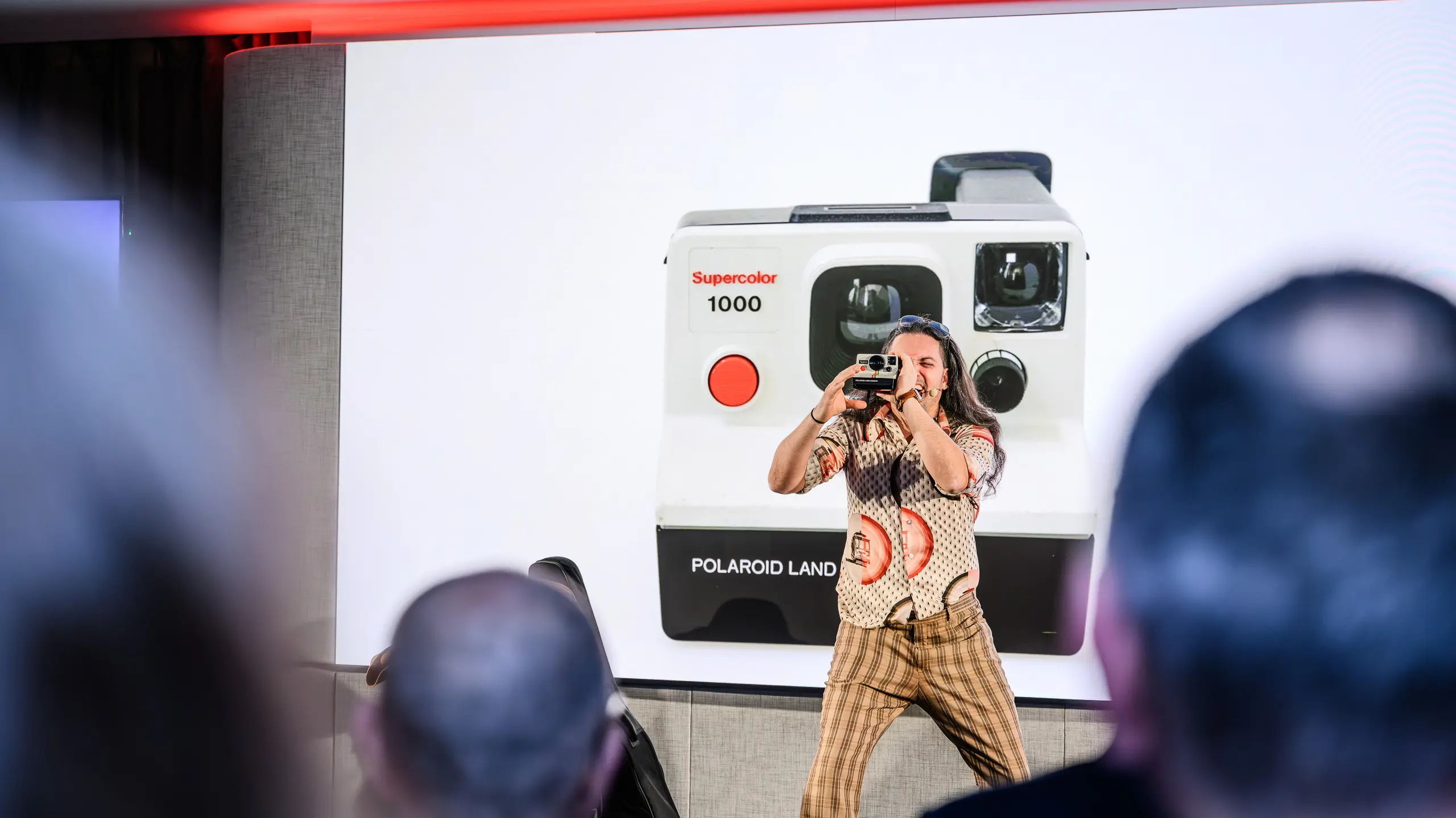 Ustvarjalni trenutek na predstavitvi PREFARENZEN 2026: zabaven voditelj z retro polaroidnim fotoaparatom ujame občinstvo – pred ogromno projekcijo ikoničnega Supercolor 1000. Umetniški prispevek gradi most med preteklostjo in prihodnostjo, tako kot PREFA s svojimi trajnimi aluminijastimi izdelki. Večer poln čustev, inovacij in estetskih impulzov.
