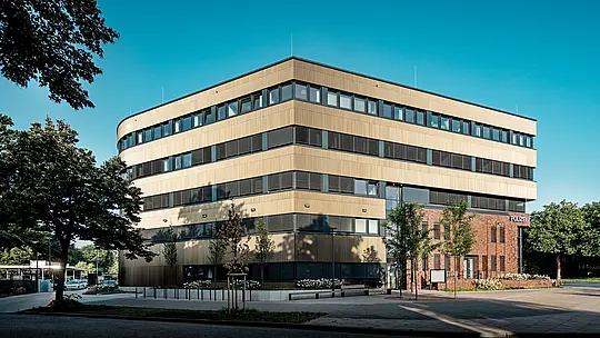 Širok pogled na zunanjost policijske postaje 43 v Hamburgu: jasno strukturirana nova stavba z zaobljenimi robovi in vodoravnimi pasovi oken navdušuje s približno 1.100 m² veliko PREFA aluminijasto fasado iz ekstrudiranih zobčastih profilov v barvi srednje bronaste. Fino strukturirana površina deluje živahno, odvisno od padanja svetlobe, in stavbi daje globino ter eleganco. V zanimivem kontrastu z obstoječo opeko nastane izrazit magnet za poglede, medtem ko PREFA aluminij prepriča s svojo dolgo življenjsko dobo, odpornostjo proti koroziji in majhnimi zahtevami po vzdrževanju.