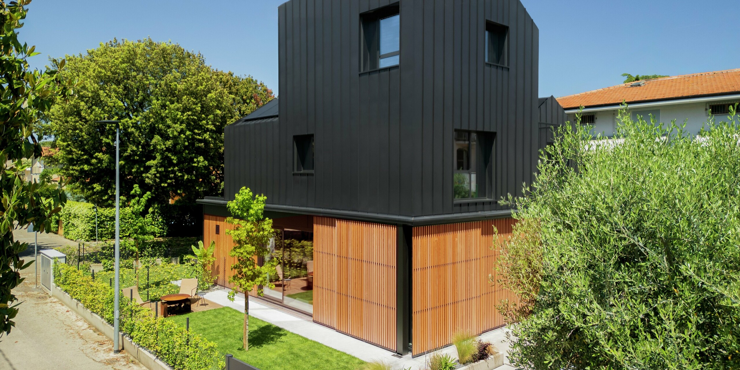 Straßenansicht eines modernen Doppelhauses in Cattolica: elegante Kombination aus Holzlamellen im Erdgeschoss und Aluminiumfassade mit PREFALZ in Schwarzgrau. Die klare Linienführung und die hochwertige PREFA Oberfläche bieten Langlebigkeit, Korrosionsschutz und zeitlose Architektur unter sonnigem Himmel.