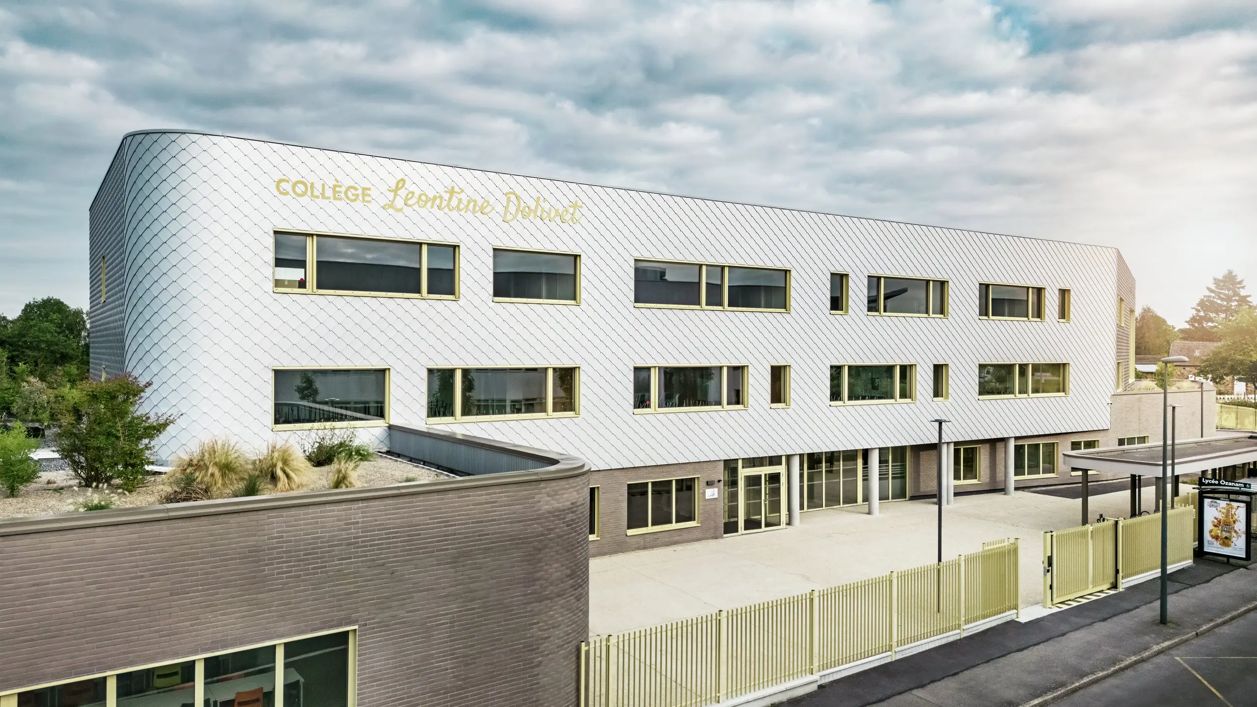Sprednji pogled na Collège Léontine Dolivet v Cesson-Sévigné z moderno aluminijasto fasado iz PREFA fasadnih rombov v kovinsko srebrni barvi. Spreminjajoča se površina daje stavbi lahkotnost, medtem ko jasne linije in velika okna zagotavljajo preglednost. Rumeni okenski okvirji dajejo živahne poudarke. V ospredju so vidni svetel predprostor, ograja in sosednja cesta. Trajna fasadna rešitev PREFA združuje dizajn, vremensko odpornost in trajnost – arhitekturni magnet z elegantnim oblikovanjem.