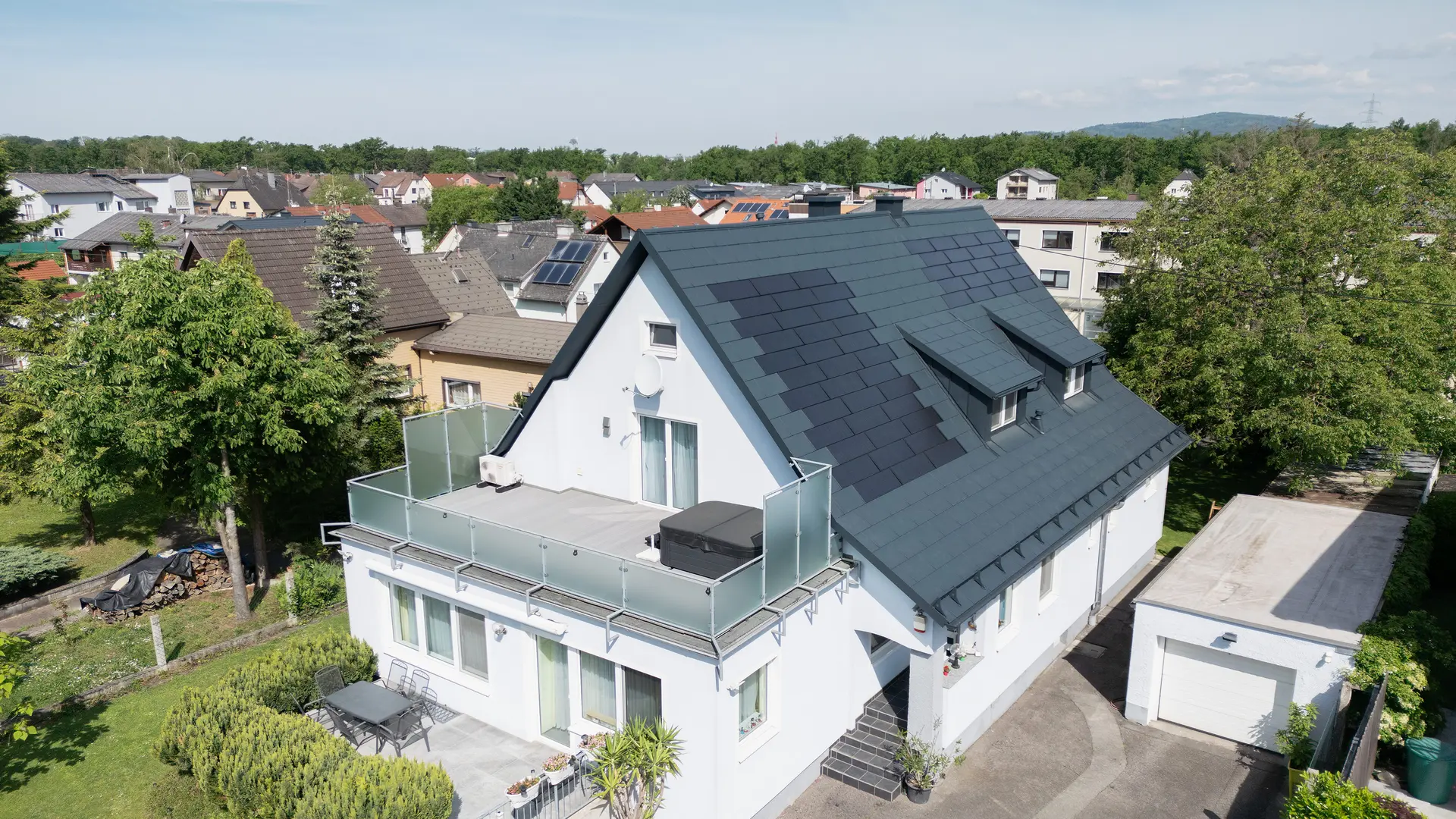 Pogled iz ptičje perspektive na enodružinsko hišo po obnovi strehe. Streha je zdaj pokrita z velikimi solarnimi strešnimi ploščami PREFA in strešnimi ploščami PREFA R.16 v barvi P.10 antracitna. Solarne strešne plošče so enakomerno razporejene in zagotavljajo moderno, trajnostno rešitev. Stavba zdaj deluje moderno in okolju prijazno. Balkon v ospredju ima stekleno ograjo in se nahaja v zgornjem nadstropju stavbe.