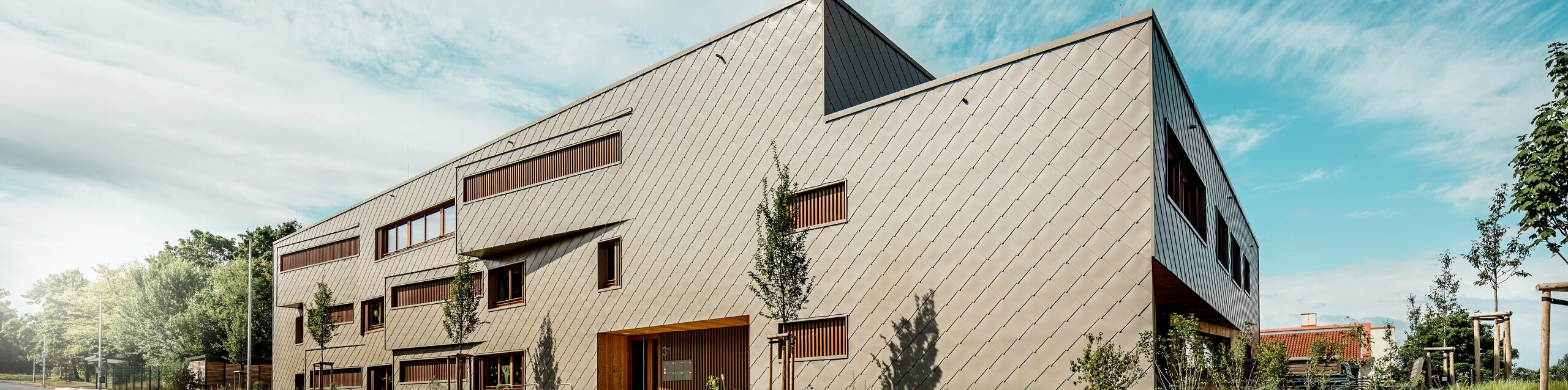 Vrtec v mestu Alzenau se s svojimi izrazitimi, dinamičnimi linijami in visokokakovostno PREFA aluminijasto fasado v bronasti barvi predstavlja iz izrazite perspektive od spodaj. Diagonalna razporeditev fasadnih rombov 44 × 44 daje stavbi jasno strukturo in elegantno, homogeno površino. Premaknjena okna in navpične lesene lamele dajejo ritmične poudarke, medtem ko zmanjšana oblika signalizira moderno zadržanost. Kombinacija trajnega aluminija, naravnega lesa in premišljenega zelenega koncepta ob cesti poudarja visoke zahteve glede trajnosti in kakovosti gradnje. Ta slika impresivno prikazuje, kako lahko funkcionalna arhitektura s PREFA izdelki prepriča z oblikovanjem in postavi merila v urbanističnem smislu.