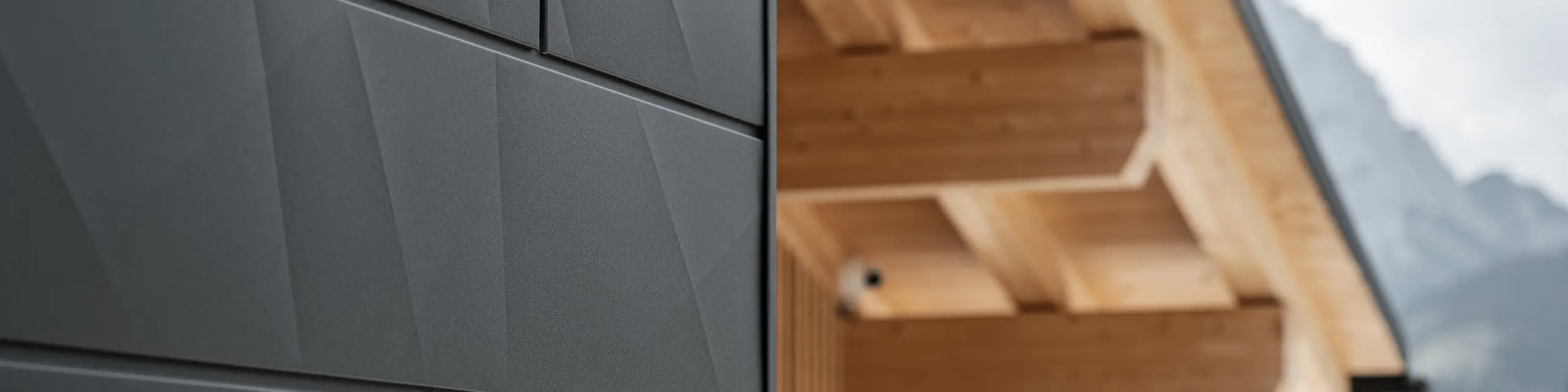 Podroben posnetek fasadnih panelov PREFA Siding.X v odtenku P.10 črna na hiši Casa Hoffe v Sappadi, Italija. Aluminijasti fasadni sistem je mojstrsko nameščen v sodobnem, abstraktnem dizajnu, ki ustvarja privlačen kontrast z vidnimi lesenimi elementi strešne konstrukcije. Inovativen fasadni sistem se brezhibno vklopi v alpsko okolje in predstavlja združitev funkcionalnosti, vzdržljivosti in estetske gradbene kulture.
