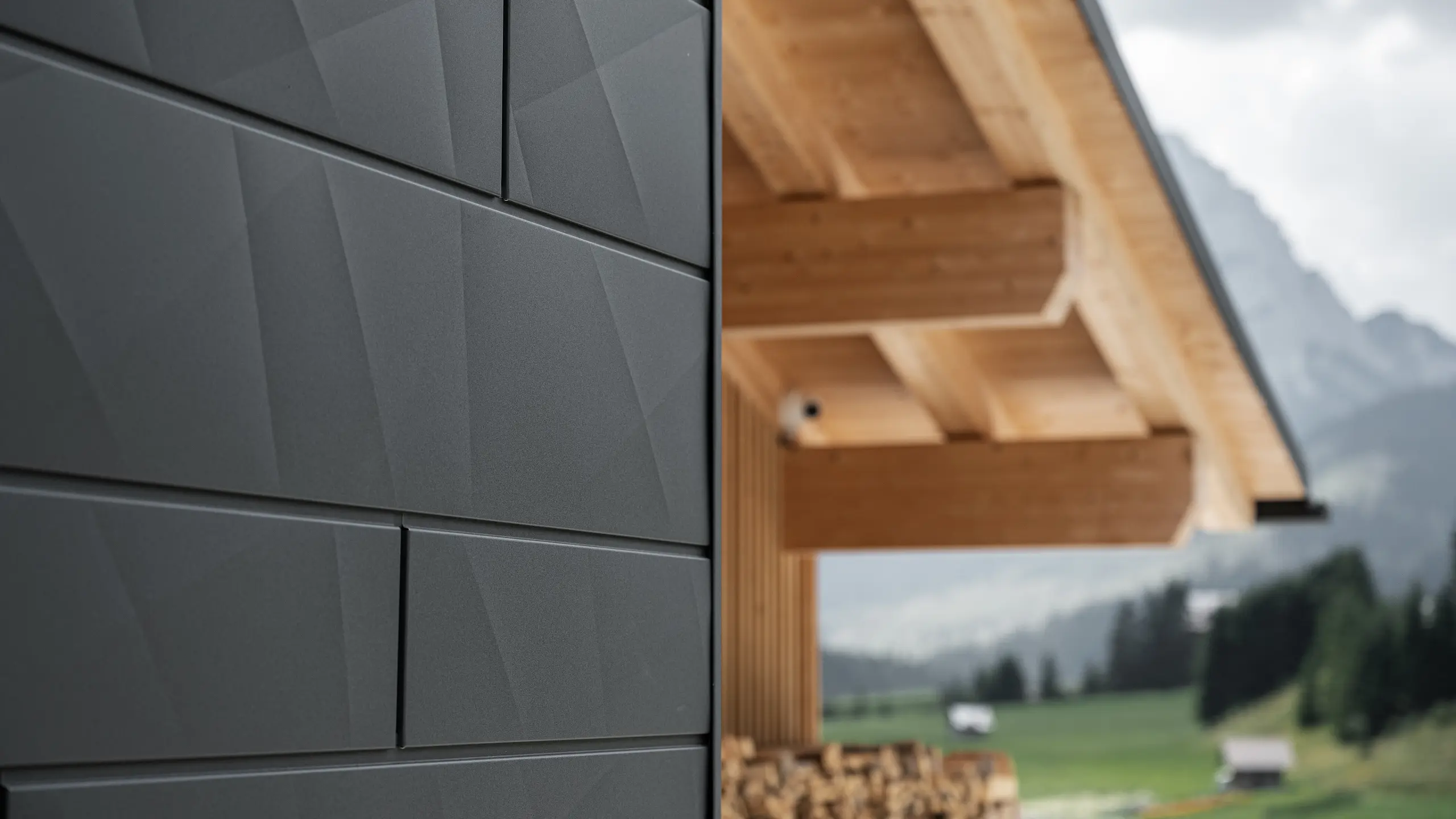Podroben posnetek fasadnih panelov PREFA Siding.X v odtenku P.10 črna na hiši Casa Hoffe v Sappadi, Italija. Aluminijasti fasadni sistem je mojstrsko nameščen v sodobnem, abstraktnem dizajnu, ki ustvarja privlačen kontrast z vidnimi lesenimi elementi strešne konstrukcije. Inovativen fasadni sistem se brezhibno vklopi v alpsko okolje in predstavlja združitev funkcionalnosti, vzdržljivosti in estetske gradbene kulture.