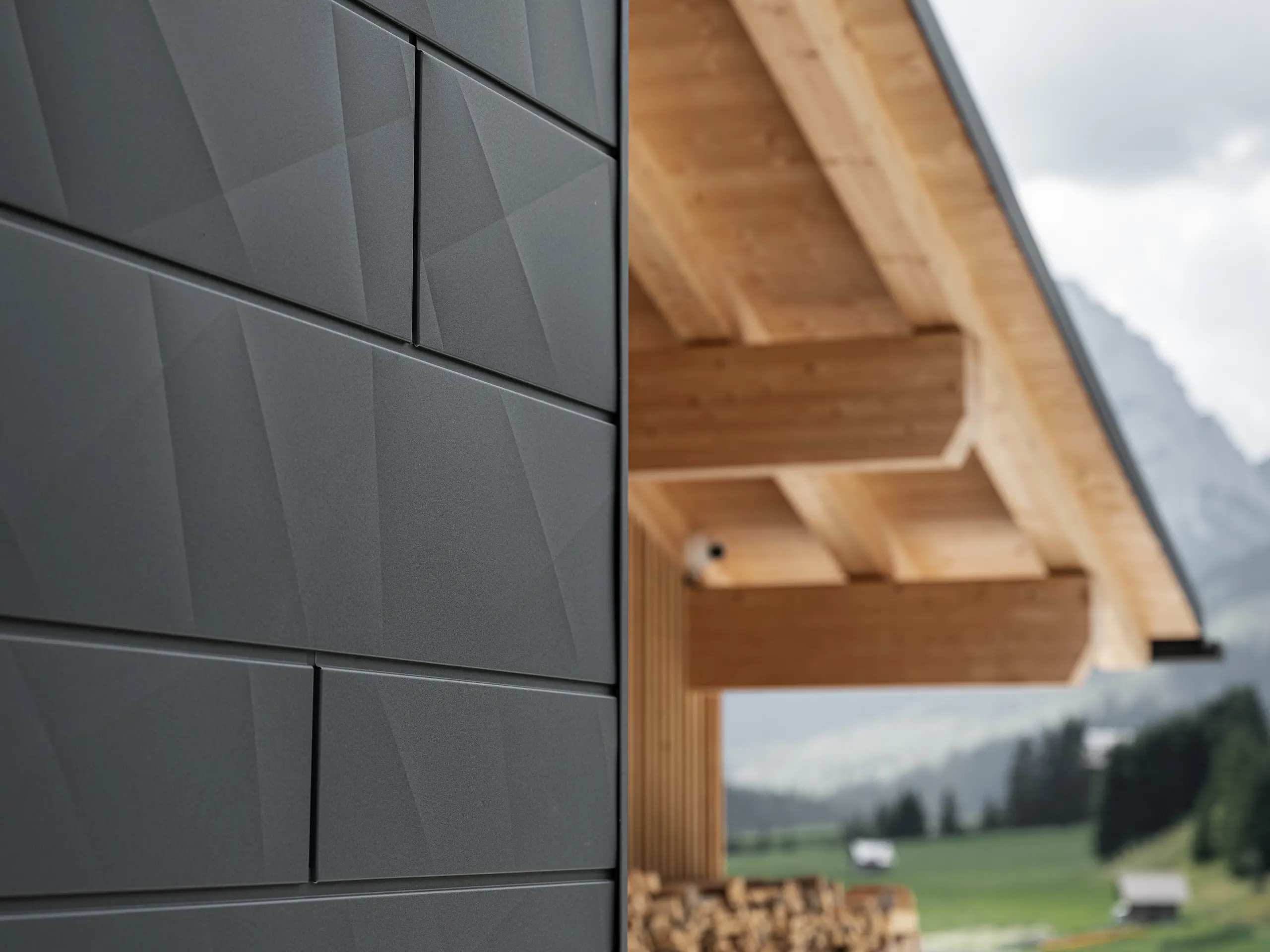 Podroben posnetek fasadnih panelov PREFA Siding.X v odtenku P.10 črna na hiši Casa Hoffe v Sappadi, Italija. Aluminijasti fasadni sistem je mojstrsko nameščen v sodobnem, abstraktnem dizajnu, ki ustvarja privlačen kontrast z vidnimi lesenimi elementi strešne konstrukcije. Inovativen fasadni sistem se brezhibno vklopi v alpsko okolje in predstavlja združitev funkcionalnosti, vzdržljivosti in estetske gradbene kulture.
