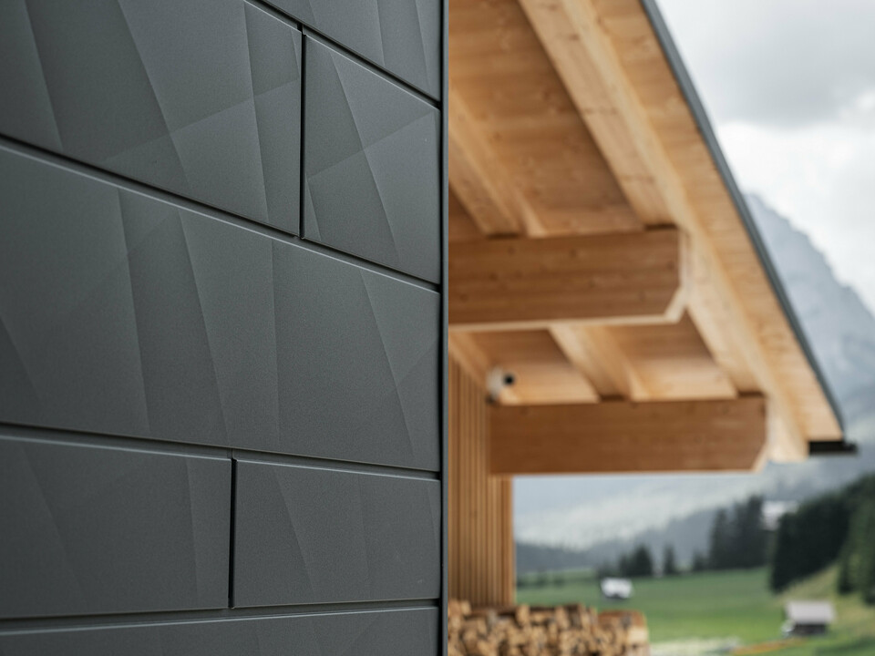 Približni pogled na fasadne plošče Siding.X v barvi P.10 črna od PREFA na hiši Casa Hoffe v Sappadi v Italiji. Fasadni sistem iz aluminija je spretno vgrajen v sodoben, abstrakten dizajn, ki ustvarja privlačen kontrast z vidnimi lesnimi elementi strešne konstrukcije. Inovativna fasadna rešitev se brezhibno vklaplja v alpsko okolje in predstavlja povezavo med funkcionalnostjo, trajnostjo in estetsko gradbeno kulturo.