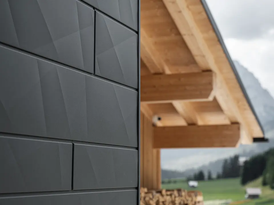 Podroben posnetek fasadnih panelov PREFA Siding.X v odtenku P.10 črna na hiši Casa Hoffe v Sappadi, Italija. Aluminijasti fasadni sistem je mojstrsko nameščen v sodobnem, abstraktnem dizajnu, ki ustvarja privlačen kontrast z vidnimi lesenimi elementi strešne konstrukcije. Inovativen fasadni sistem se brezhibno vklopi v alpsko okolje in predstavlja združitev funkcionalnosti, vzdržljivosti in estetske gradbene kulture.