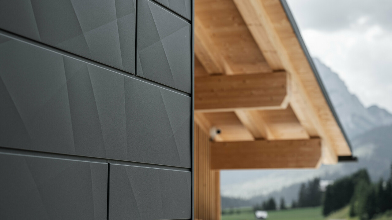 Podroben posnetek fasadnih panelov PREFA Siding.X v odtenku P.10 črna na hiši Casa Hoffe v Sappadi, Italija. Aluminijasti fasadni sistem je mojstrsko nameščen v sodobnem, abstraktnem dizajnu, ki ustvarja privlačen kontrast z vidnimi lesenimi elementi strešne konstrukcije. Inovativen fasadni sistem se brezhibno vklopi v alpsko okolje in predstavlja združitev funkcionalnosti, vzdržljivosti in estetske gradbene kulture.