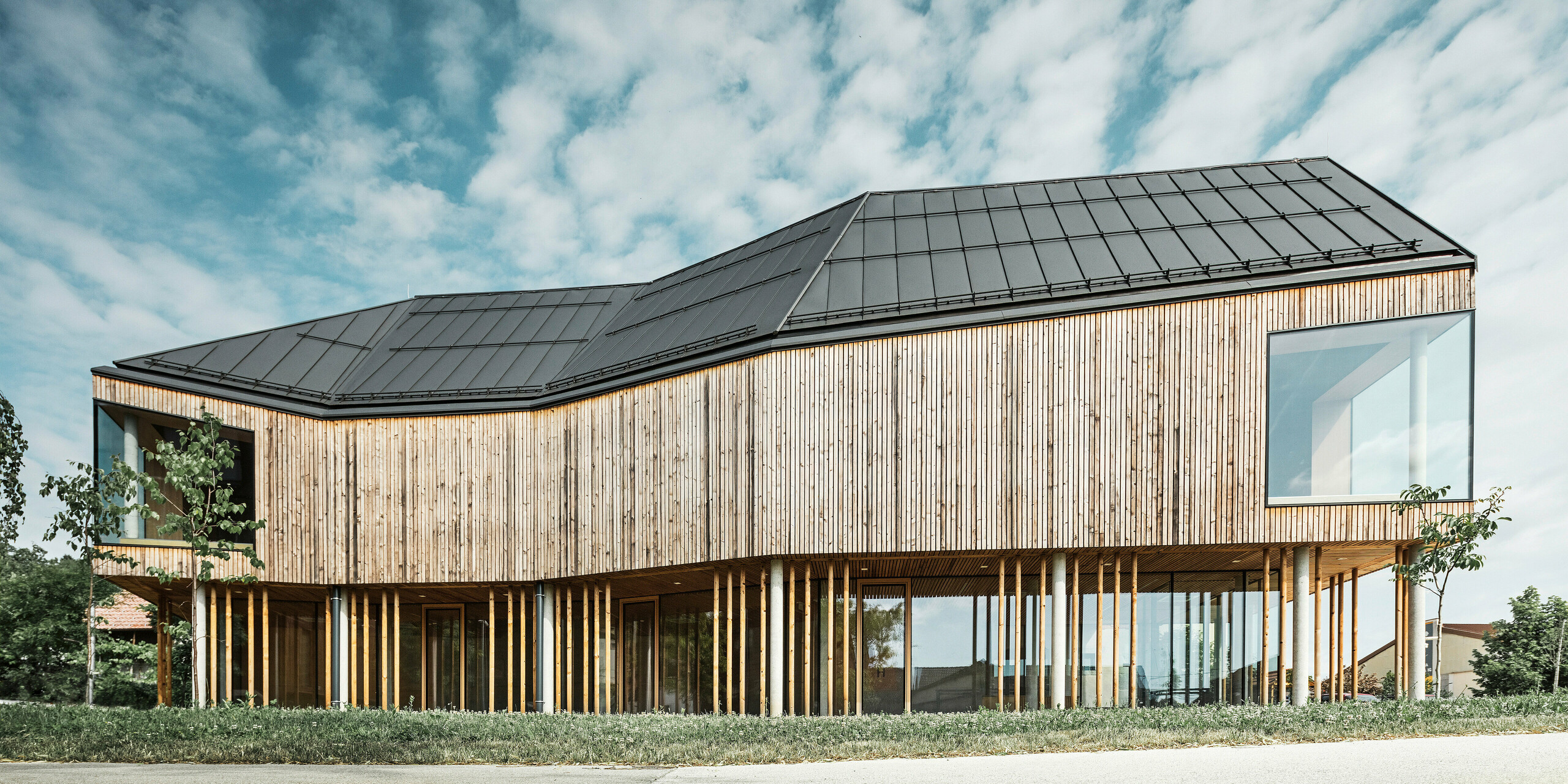 Pogled s strani na ekološko zasnovan center za obiskovalce na Igu v Sloveniji s temno sivo streho PREFALZ, ki poudarja čiste linije in sodoben arhitekturni jezik stavbe. Interakcija naravne lesene obloge in obsežne zasteklitve, ki odseva modro nebo in oblake, dokazuje povezavo med funkcionalnostjo in estetiko. Dizajn se brezhibno zliva s podeželskim okoljem in odraža inovativne metode gradnje.