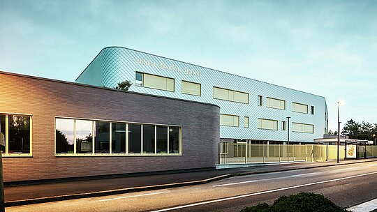 Večerni pogled na Collège Léontine Dolivet v Cesson-Sévigné. V ospredju poteka jasno označena cesta, ob njej pa je nizka stavba z velikimi steklenimi okni. Za njo se dviga glavna stavba z ukrivljeno aluminijasto fasado iz PREFA fasadnih rombov v kovinskosrebrni barvi, ki se spreminja v mehki svetlobi. Več pravokotnih oken in svetla ograja strukturirajo pogled. Prizor osvetljujejo ulične svetilke, nebo je hladno modro – sodoben, eleganten pogled.