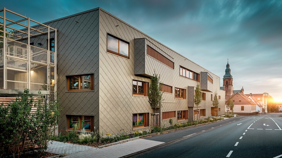  V nežni svetlobi sončnega zahoda se vrtec v Alzenauu pokaže v svoji najbolj čarobni podobi. Fasada iz bronastih PREFA fasadnih rombov 44 × 44 odseva večerno svetlobo in poudarja visokokakovostno, strukturirano površino moderne hibridne stavbe. Jasno strukturirana arhitektura z zamaknjenimi okenskimi trakovi in navpičnimi lesenimi lamelami je v zanimivem kontrastu z zgodovinsko cerkvijo v ozadju. Na levi strani slike filigranska kovinska konstrukcija zagotavlja rahlo preglednost zunanjega stopnišča. Urejena zelena površina ob stavbi poudarja ekološko naravnanost. Ta slika na impresiven način združuje moderno gradbeno kulturo, trajnostno PREFA aluminijasto tehnologijo in harmonično vključevanje v zgodovinsko mestno podobo Alzenaua.