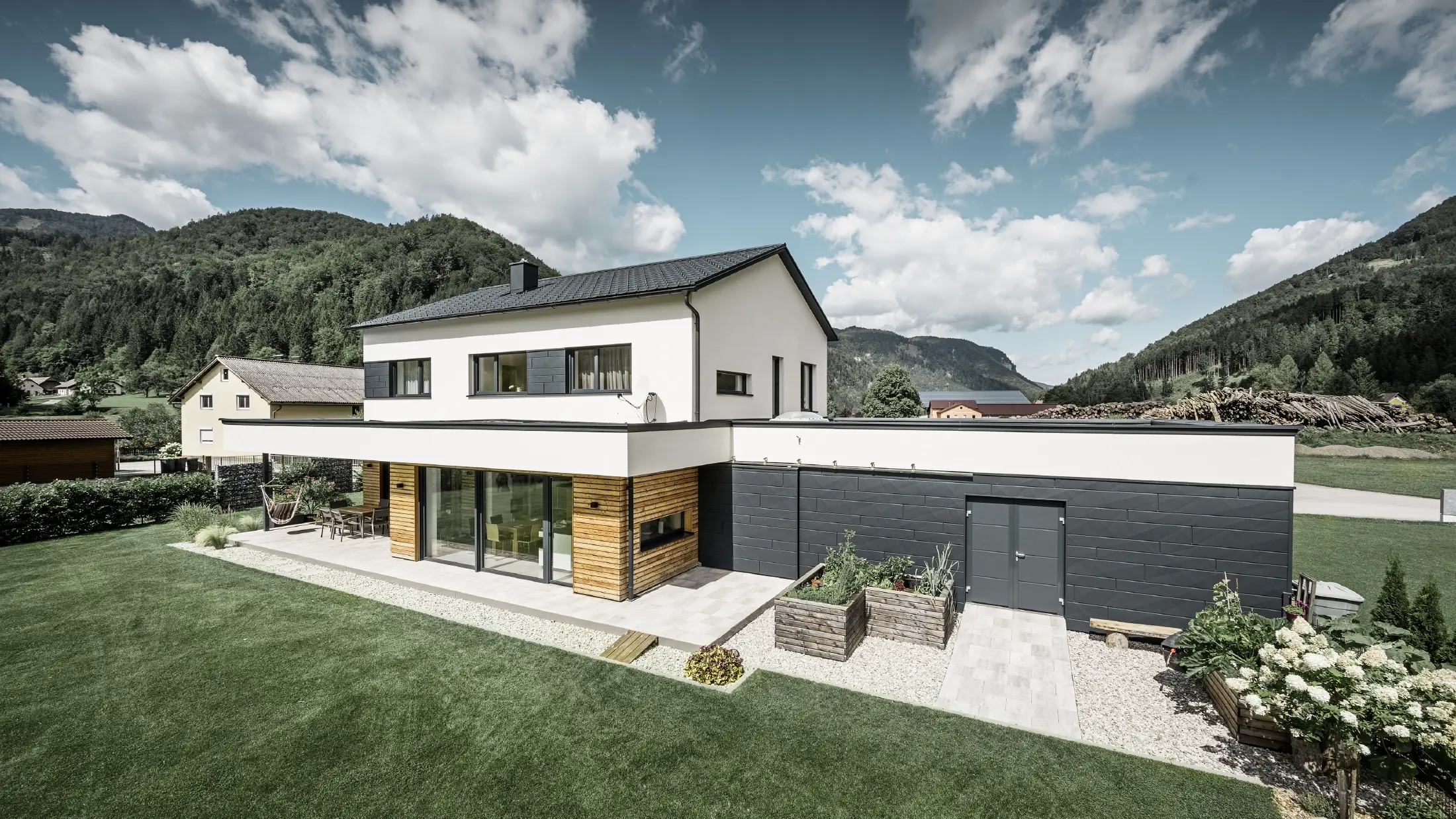 Sodobna samostojna hiša z belim ometom in PREFA Siding.X v antracitni barvi P.10, obkrožena z idiličnim podeželjem. Temna fasada je v kontrastu s svetlo okolico. Strešne plošče PREFA in strešno odvodnjavanje dopolnjujejo trajno zasnovo.