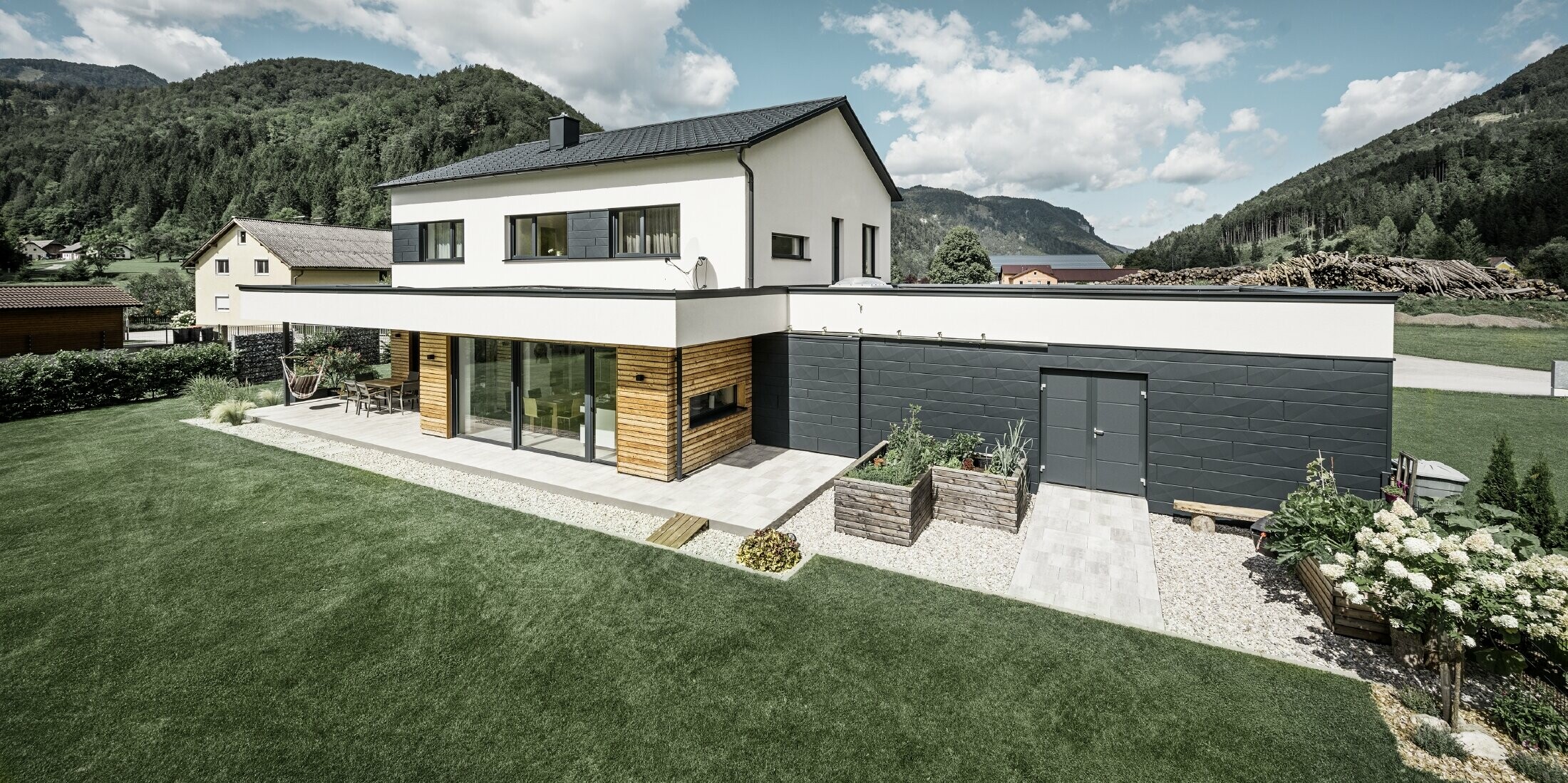 Das Bild zeigt ein modernes Einfamilienhaus in einer idyllischen Landschaft mit einer klaren Sicht auf bewaldete Hügel im Hintergrund. Das Haus zeichnet sich durch eine Kombination aus weißen Putzwänden und einer markanten Fassadenverkleidung mit PREFA Siding.X in P.10 Anthrazit aus, die für einen modernen und langlebigen Look sorgt. Die dunkle Färbung der Fassadenelemente bildet einen ansprechenden Kontrast zum hellen Außendesign und zur natürlichen Umgebung. Die Anordnung der Siding.X Fassadenpaneele ist präzise und verleiht der Immobilie einen strukturierten und hochwertigen Charakter, der typisch für PREFA Produkte ist. Das Dach des Gebäudes wurde mit der bewährten PREFA Dachplatte eingedeckt. Die Dachentwässerung erfolgt durch das Ablaufrohr und die Hängerinne von PREFA.