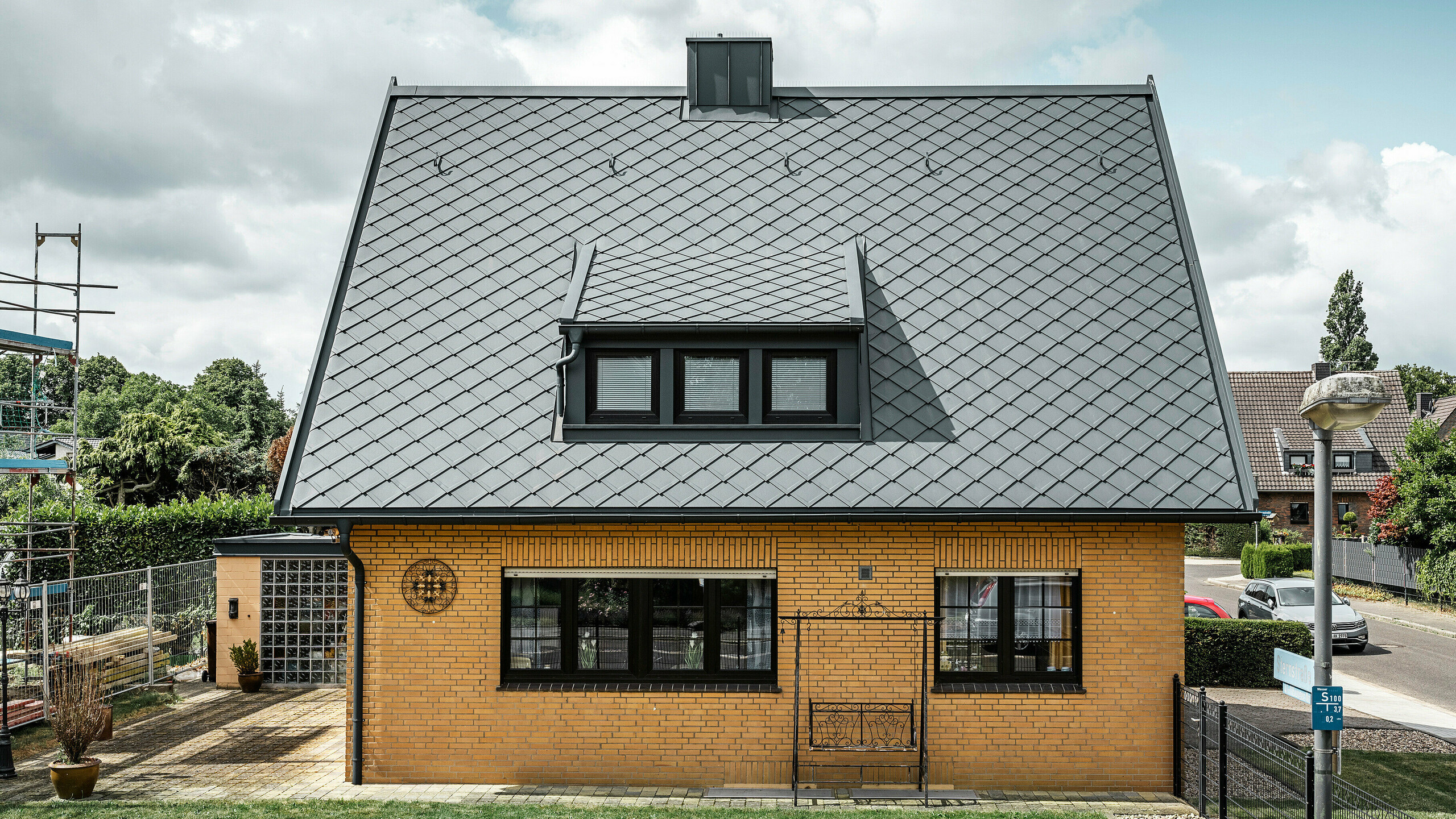 Stranski pogled na enodružinsko hišo v Tönisvorstu s PREFA streho antracitne barve s strešnimi rombi 29×29. Kombinacija tradicionalne rumene opeke s sodobnimi strešnimi elementi in dodatki iz aluminija ustvarja harmoničen videz. Mansarde in dimnik, tudi v antracitni barvi, zaokrožujejo estetsko in funkcionalno streho v malem mestu.