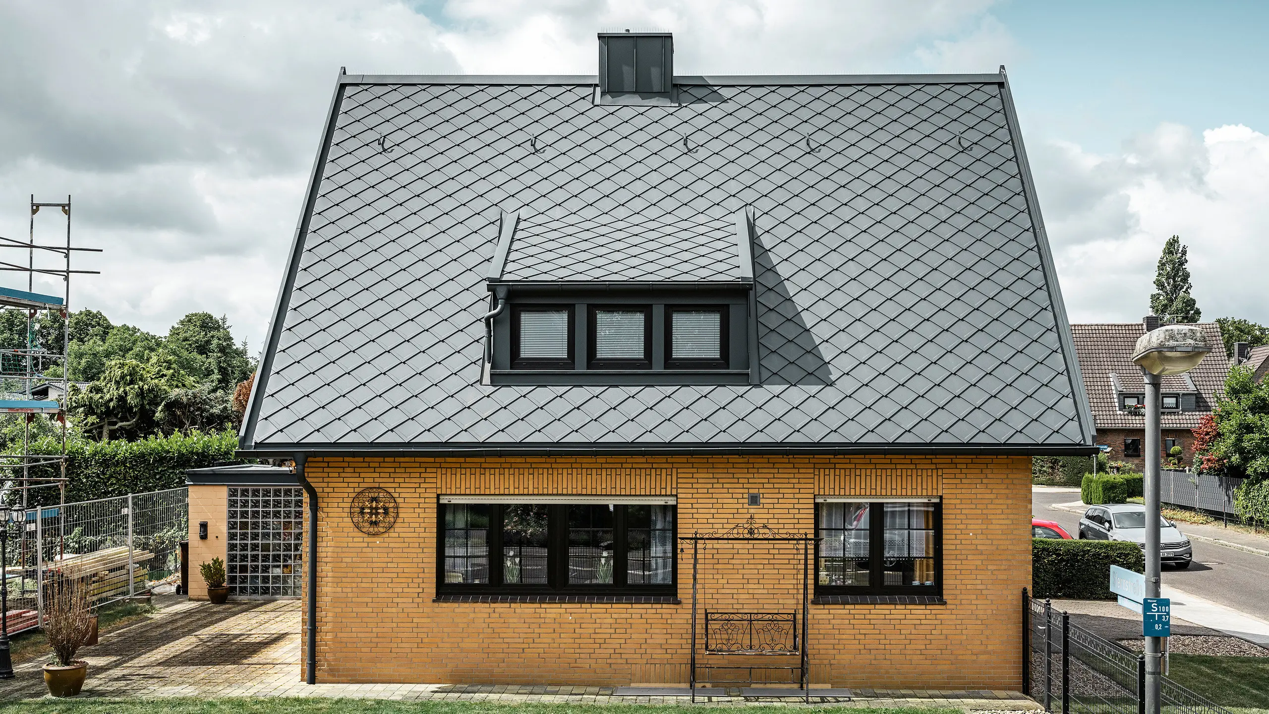Stranski pogled na enodružinsko hišo v Tönisvorstu s PREFA streho antracitne barve s strešnimi rombi 29×29. Kombinacija tradicionalne rumene opeke s sodobnimi strešnimi elementi in dodatki iz aluminija ustvarja harmoničen videz. Mansarde in dimnik, tudi v antracitni barvi, zaokrožujejo estetsko in funkcionalno streho v malem mestu.