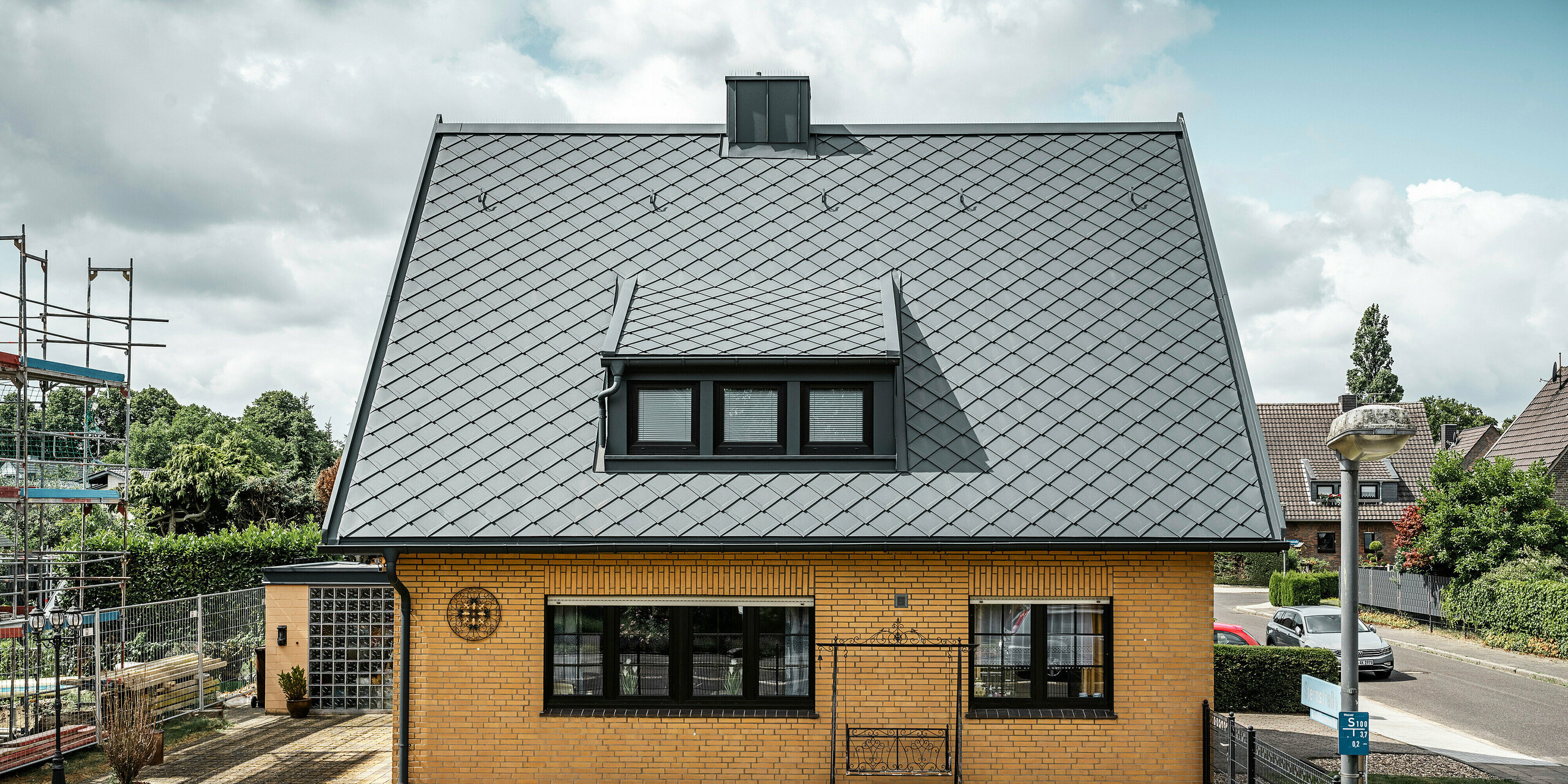 Stranski pogled na enodružinsko hišo v Tönisvorstu s PREFA streho antracitne barve s strešnimi rombi 29×29. Kombinacija tradicionalne rumene opeke s sodobnimi strešnimi elementi in dodatki iz aluminija ustvarja harmoničen videz. Mansarde in dimnik, tudi v antracitni barvi, zaokrožujejo estetsko in funkcionalno streho v malem mestu.