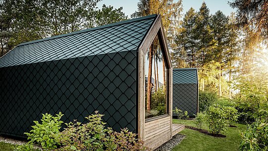 Minimalistična hišica »Tiny House« v Secret Koi Garden ob jezeru Natterer See je obdana z zelenjem in obdana s toplim večernim svetlobom. Streha in fasada sta v celoti obloženi z majhnimi PREFA strešnimi in fasadnimi rombi iz aluminija v barvi P.10 antracit, katerih natančna struktura, podobna ribji koži, ustvarja izrazite poudarke. Trajen in vremensko odporen PREFA aluminij zanesljivo ščiti arhitekturo in se odlično ujema z lesom, steklom in okoliško naravo.