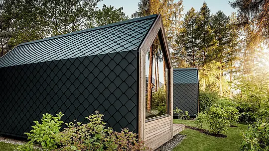 Minimalistična hišica »Tiny House« v Secret Koi Garden ob jezeru Natterer See je obdana z zelenjem in obdana s toplim večernim svetlobom. Streha in fasada sta v celoti obloženi z majhnimi PREFA strešnimi in fasadnimi rombi iz aluminija v barvi P.10 antracit, katerih natančna struktura, podobna ribji koži, ustvarja izrazite poudarke. Trajen in vremensko odporen PREFA aluminij zanesljivo ščiti arhitekturo in se odlično ujema z lesom, steklom in okoliško naravo.