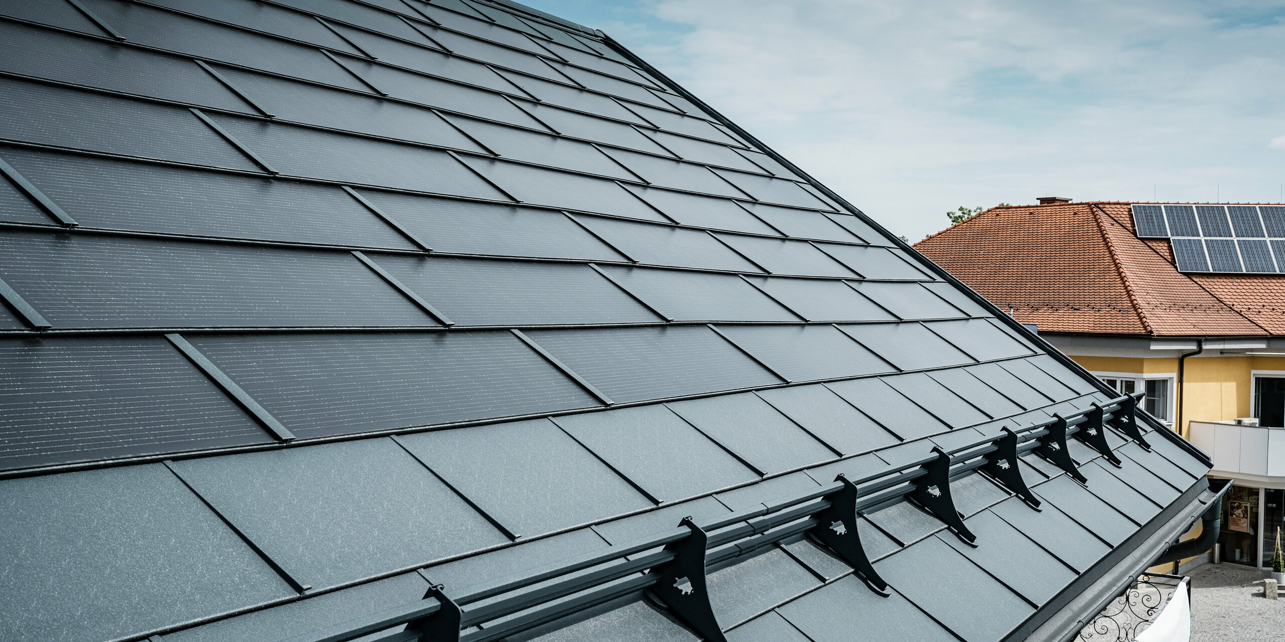 Pogled na moderno streho s PREFA solarnimi strešnimi ploščami antracitne barve. Pri napušču je bila streha opremljena s klasičnimi PREFA strešnimi ploščami R.16 in PREFA snežnim sistemom. V ozadju je mogoče videti streho sosednje hiše s tradicionalnimi strešniki in sončnimi kolektorji na strehi, kar poudarja kontrast med novo in tradicionalno zasnovo strehe. Inovativna PREFA solarna strešna plošča združuje estetiko in funkcionalnost ter ponuja trajnostno energetsko rešitev za vse vrste stavb.
