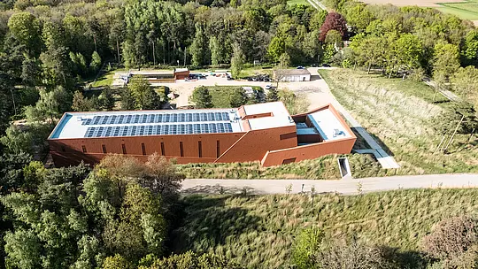 Letalski posnetek črpalne postaje za pitno vodo WML v Lottumu kot izrazitega infrastrukturnega objekta, ki se harmonično vključuje v zeleno pokrajino gozdov in travnikov. Podolgovat objekt z izrazito aluminijasto fasado PREFA Siding.X v topli rjavi barvi deluje kot skulpturalna znamenitost v okolici. Oči pritegnejo jasno strukturirana oblika stavbe, vgrajeni fotovoltaični moduli na strehi ter fino fasetirana površina fasade. Trajni, lahki in reciklirani aluminijasti paneli PREFA prepričajo z visoko odpornostjo proti vremenskim vplivom.