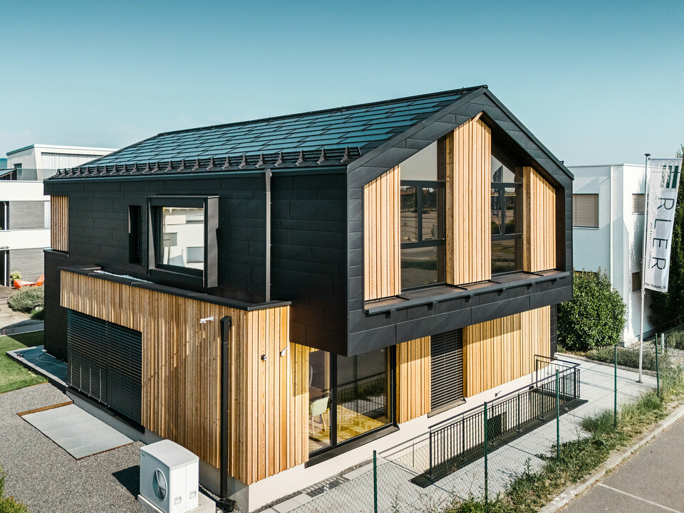 Sprednja stran vzorčne hiše Autark v centru montažnih hiš Blaue Lagune v Vösendorfu (Avstrija). Streha je prekrita s črnimi solarnimi strešnimi ploščami PREFA in strešnimi ploščami R.16 z visoko kakovostnim premazom P.10. Fasada združuje navpične lesene elemente in panele PREFA Siding.X v barvi P.10 črna, kar stavbi daje sodoben in trajnosten značaj. Na desnem robu slike plapola zastava podjetja Erler Bau GmbH.